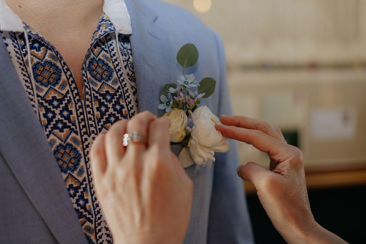 Hands pinning a white rose boutonniere on a groom wearing a blue embroidered Ukrainian wedding shirt.