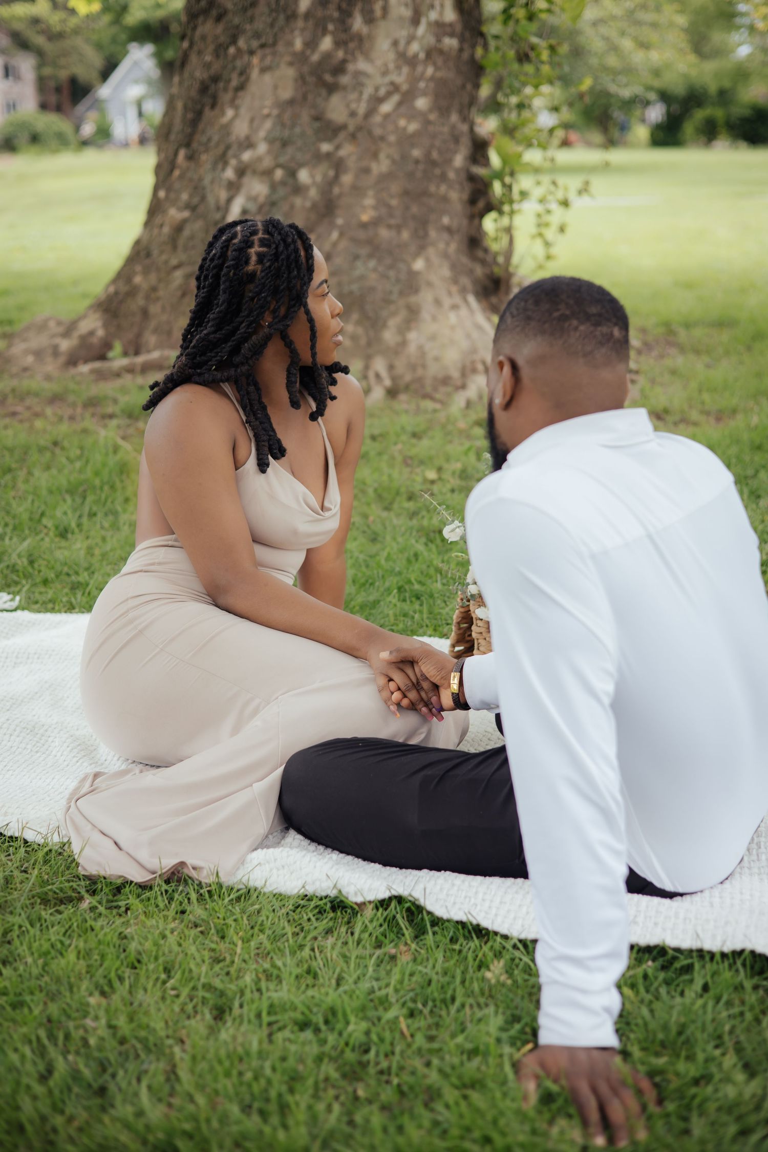 Engaged Black couple sitting on white blanket under tree, holding hands and sharing an intimate moment.