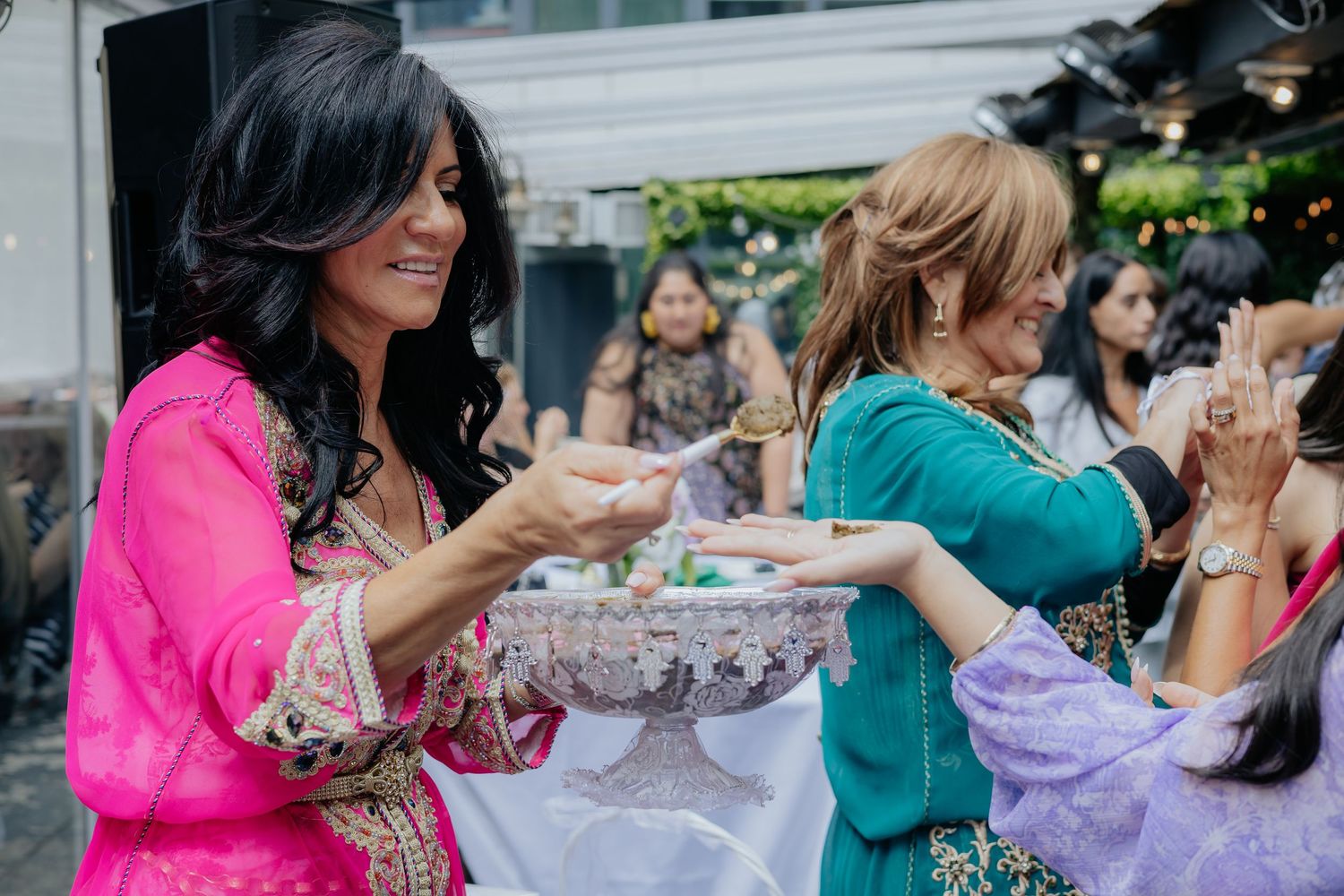 Women in colorful traditional outfits share food at a festive cultural celebration outdoors.