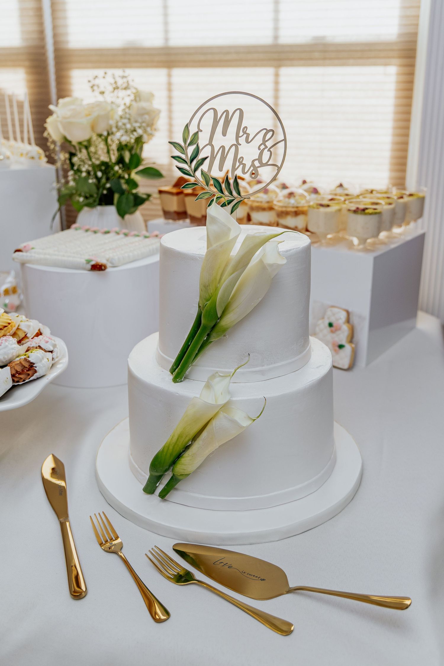 Two-tier white wedding cake with calla lily decorations and Mr & Mrs cake topper on elegant table.
