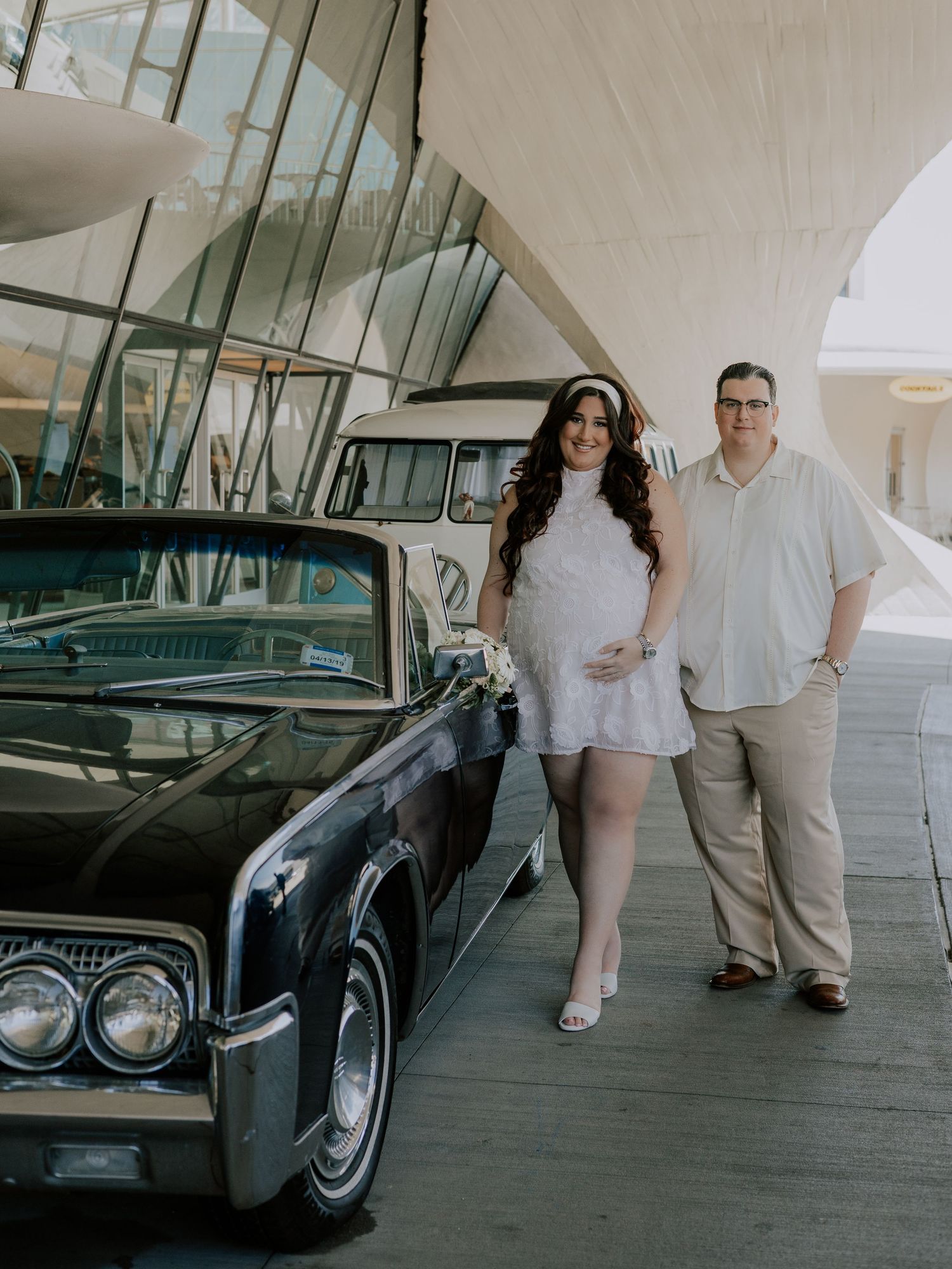 Pregnant bride and groom pose beside vintage black Lincoln Continental at TWA Hotel entrance.