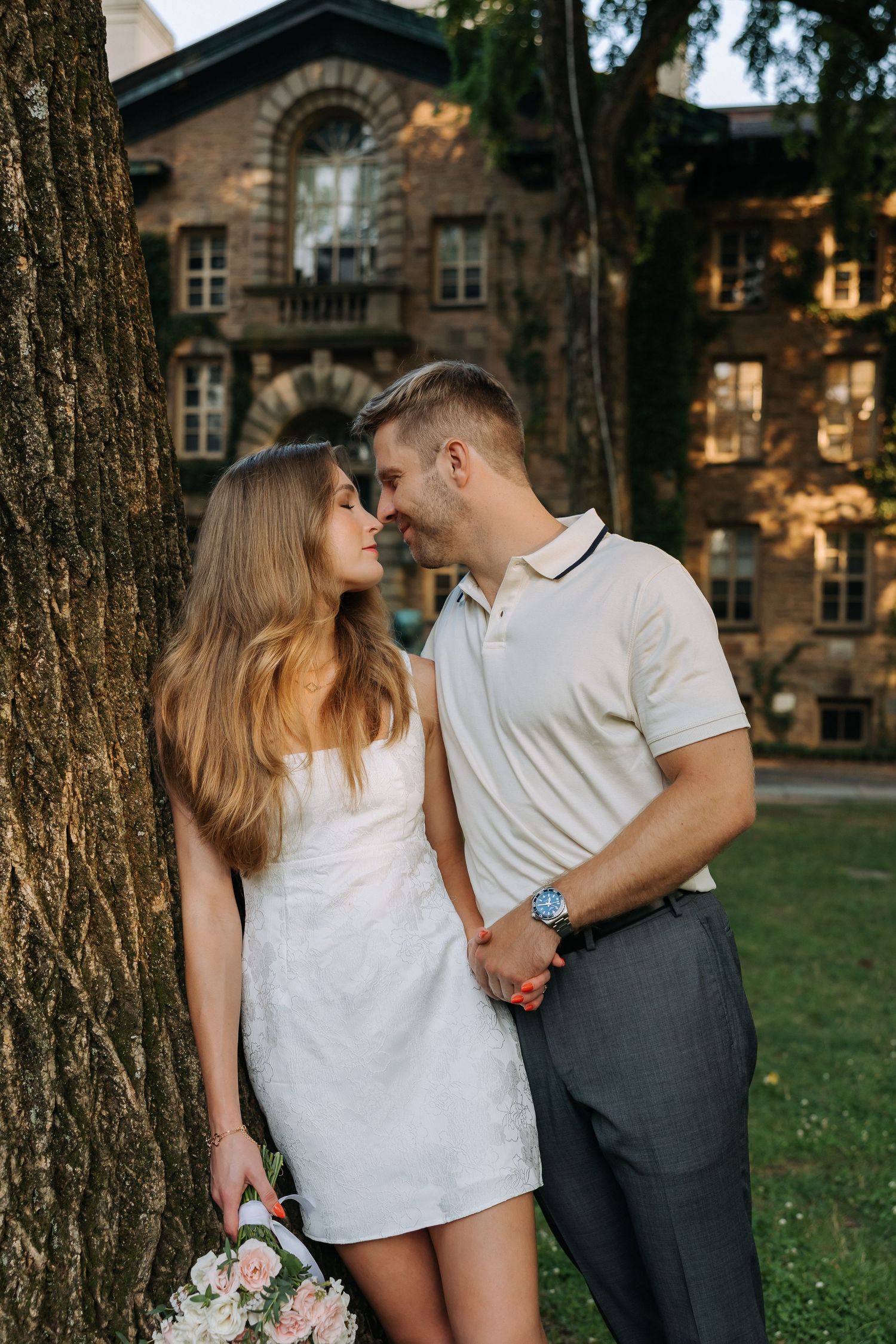A couple in love stands by a large tree, woman in white dress holding flowers, man in polo shirt, gazing at each other.