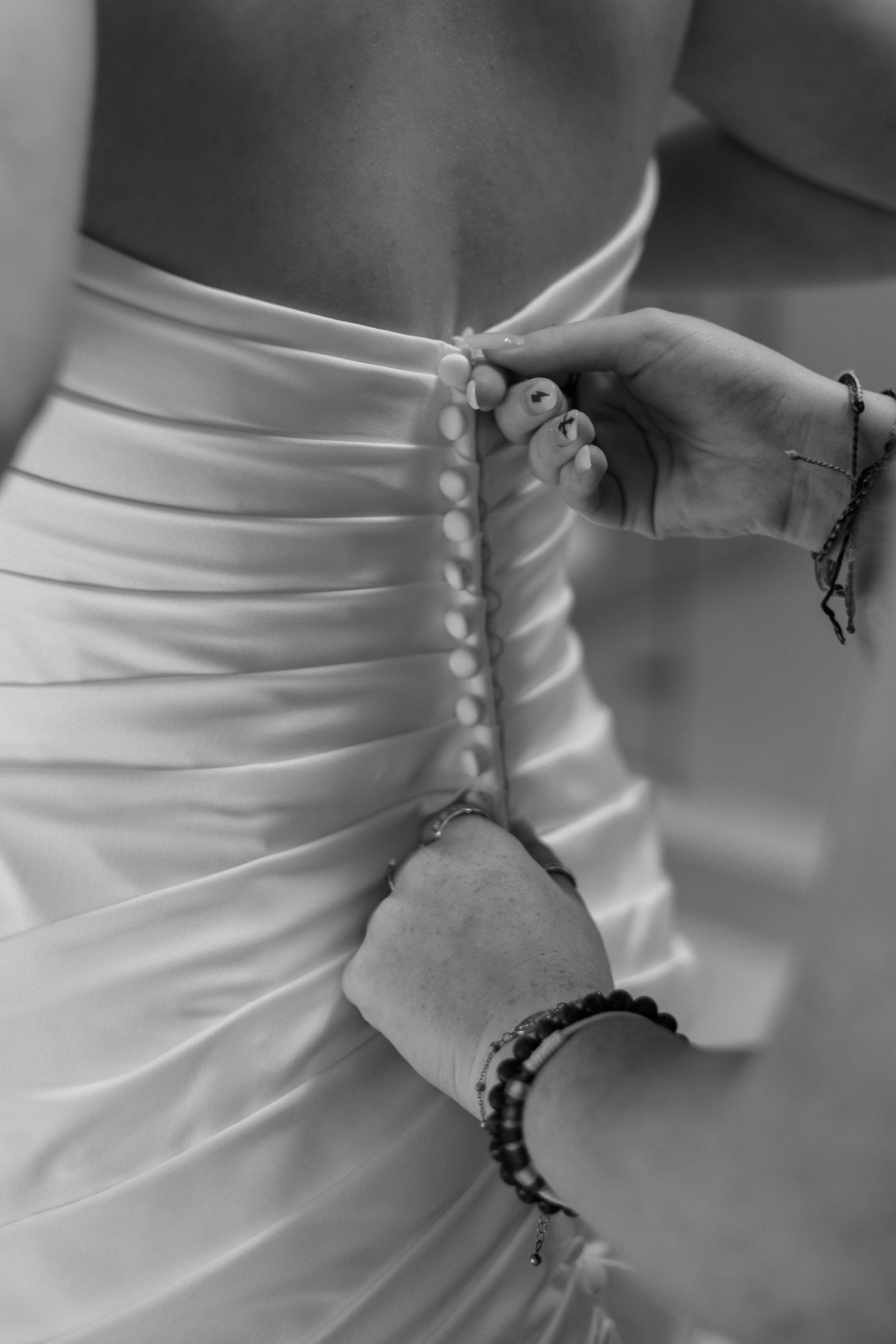 Black and white close-up of hands fastening pearl buttons on back of wedding gown.