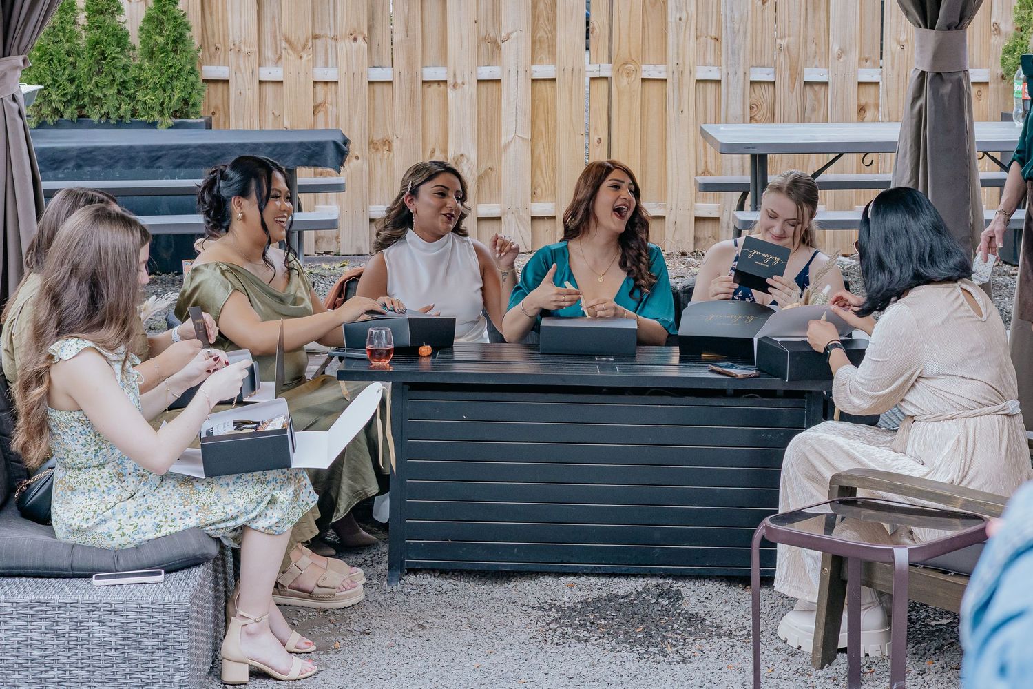 Excited group of women reacting to bridesmaid proposal boxes at outdoor patio gathering.