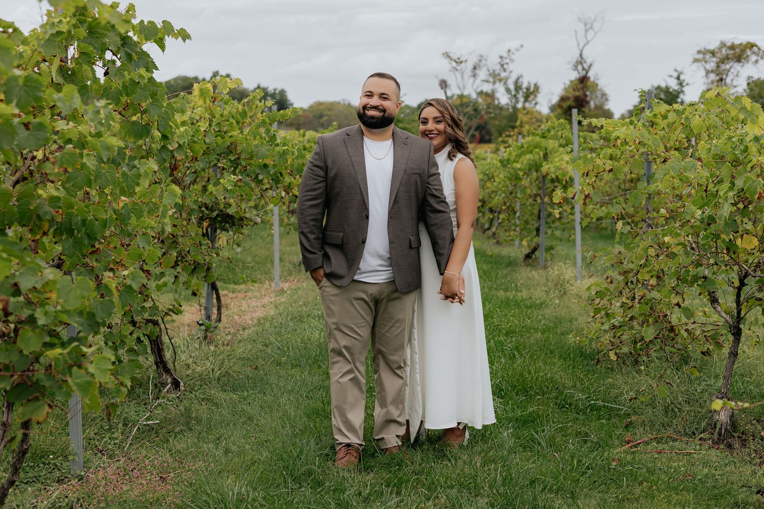 Couple poses hand-in-hand in a lush vineyard, bride in white dress and groom in a brown blazer.