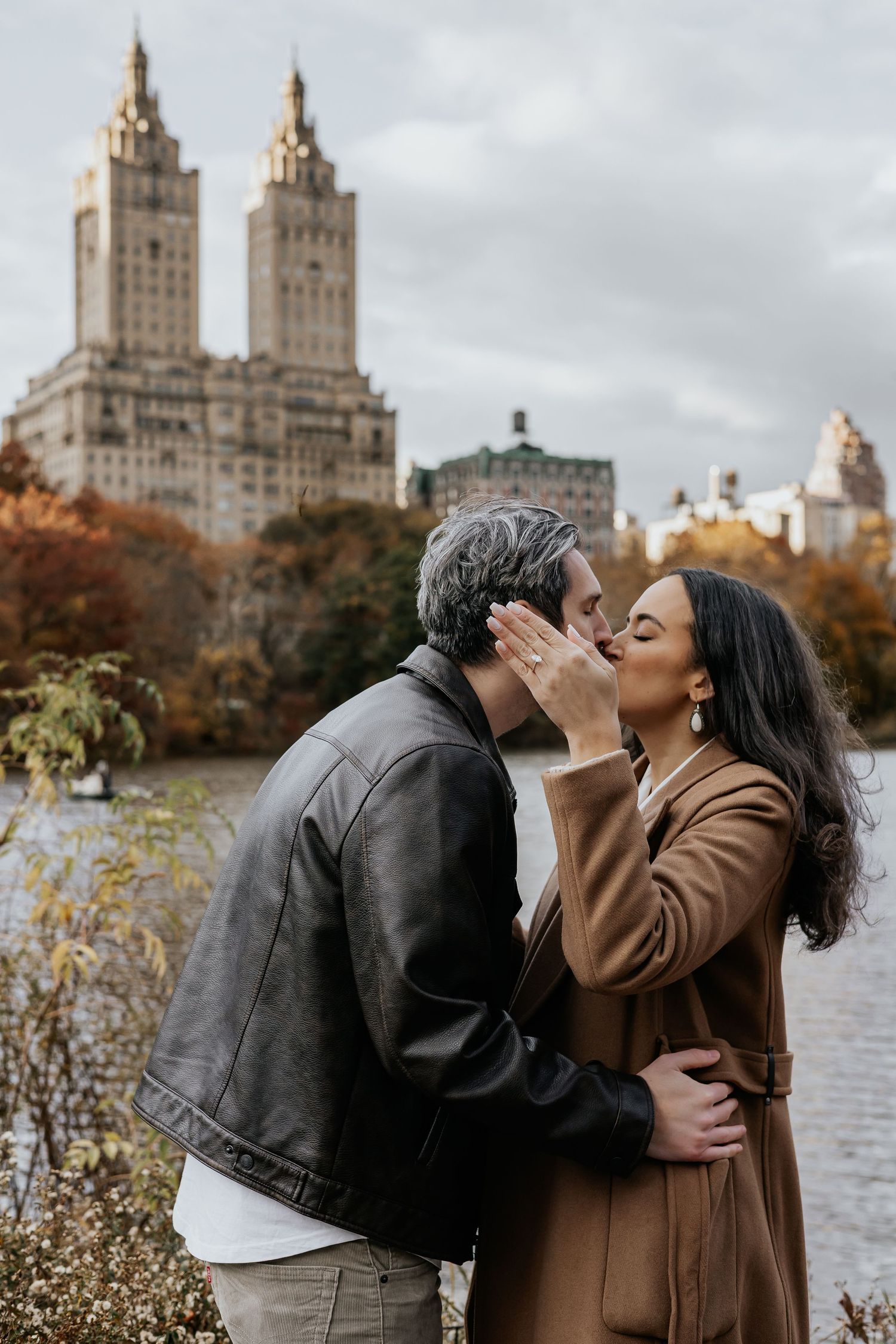 Couple sharing a romantic kiss near Central Park lake with iconic NYC skyline in fall foliage.