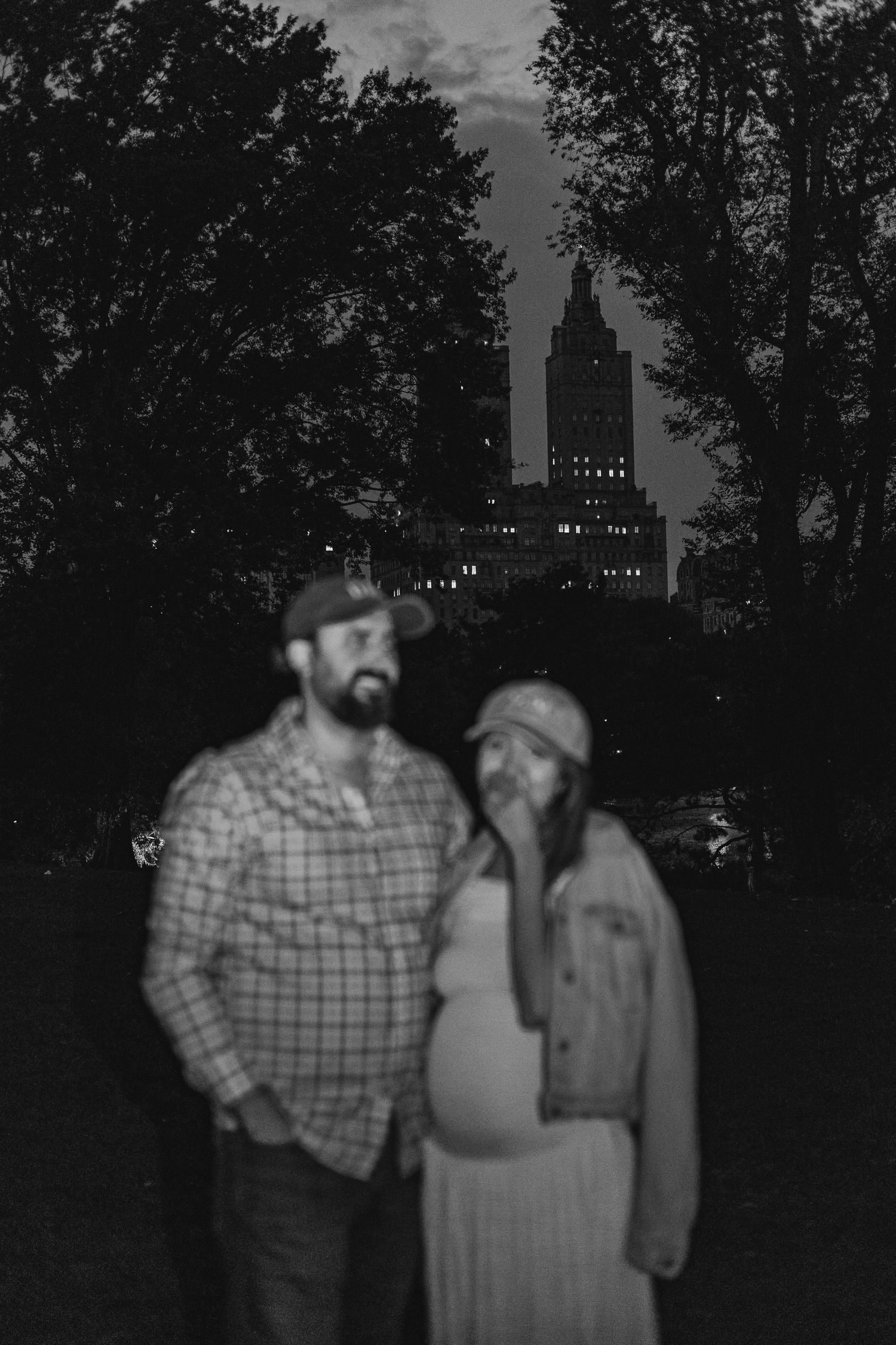 Black and white moody maternity photo of couple with NYC skyline at night.