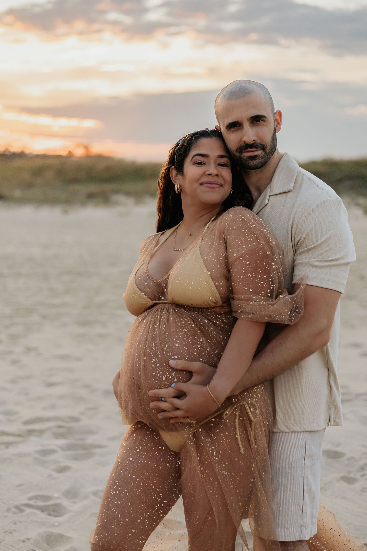 Couple posing on sandy beach at sunset, man holding pregnant partners belly in gold sheer dress.