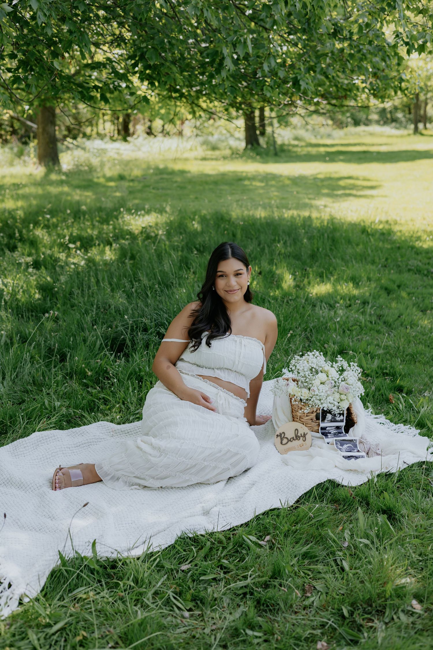 Pregnant woman in white two-piece outfit sits on blanket in lush green park with baby ultrasound photos and flower basket.