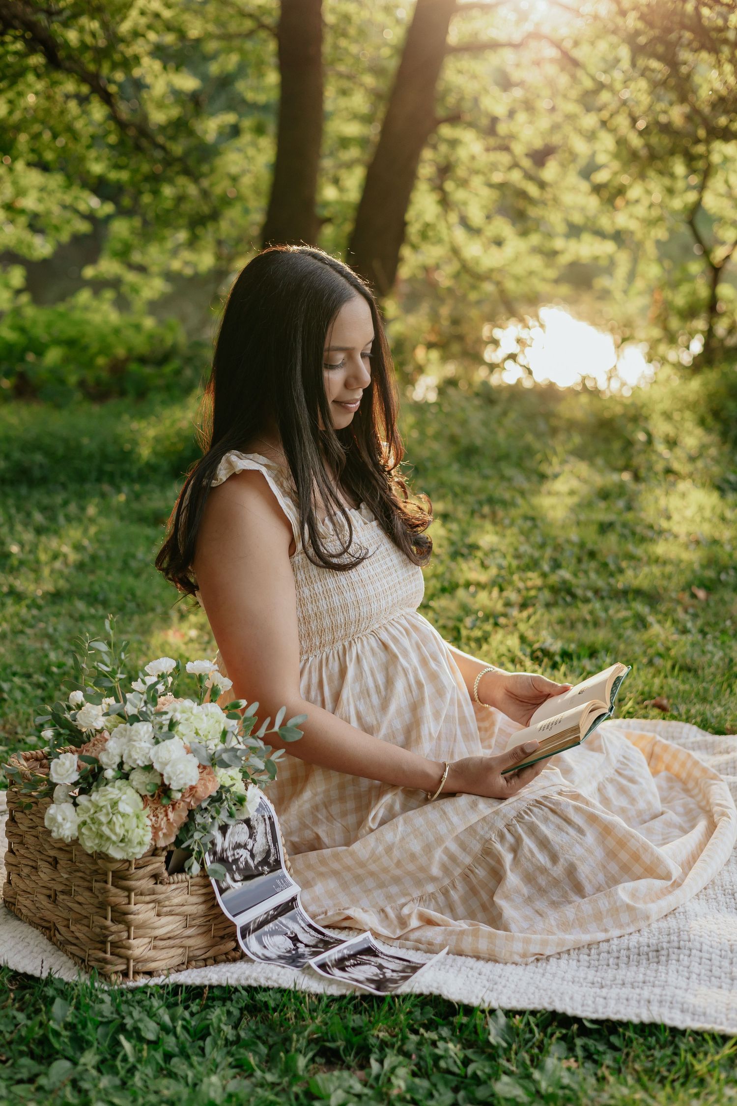 Pregnant woman in gingham dress reading book beside flower basket in sunlit park.