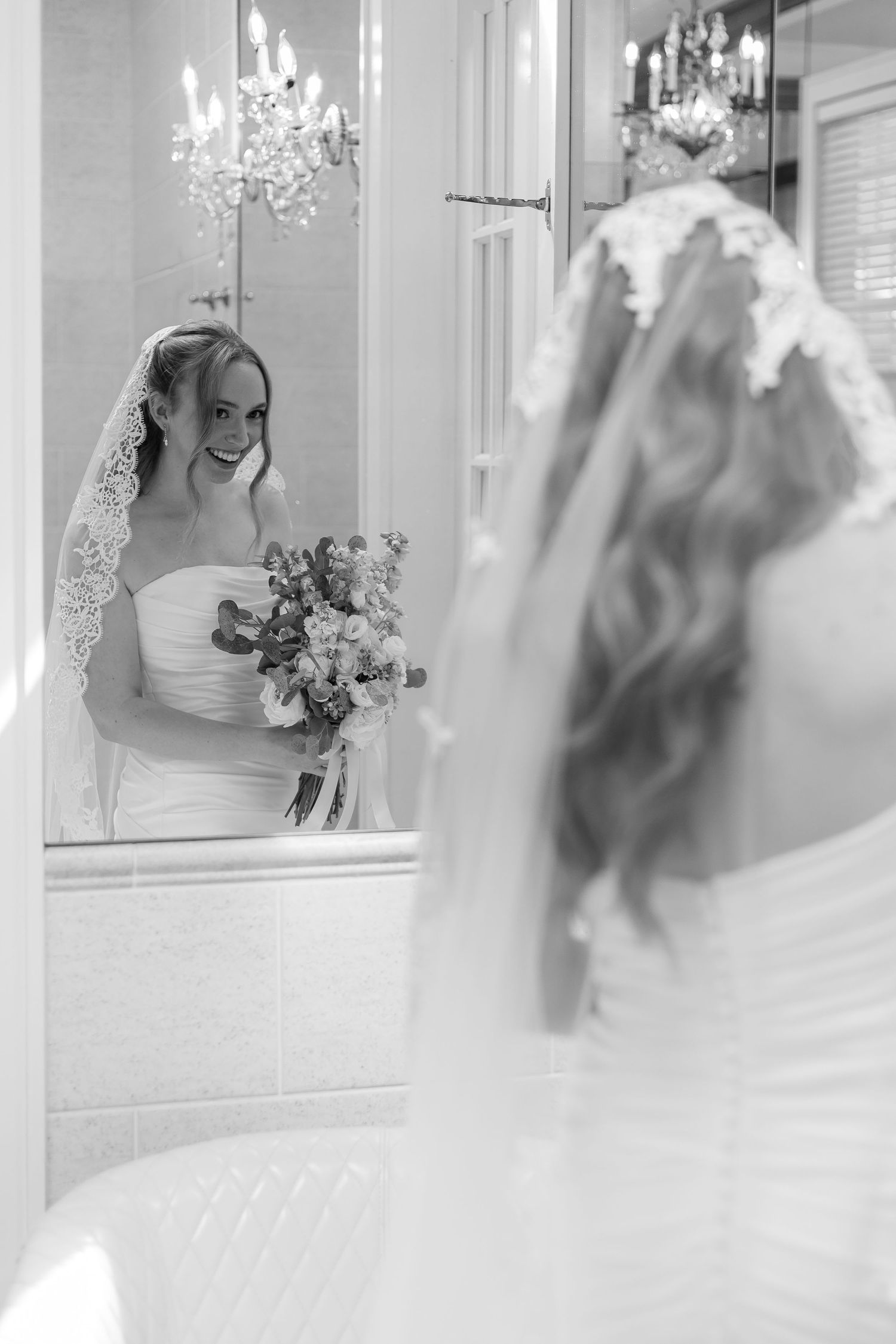 Black and white photo of bride in lace veil smiling at her reflection holding a wedding bouquet.
