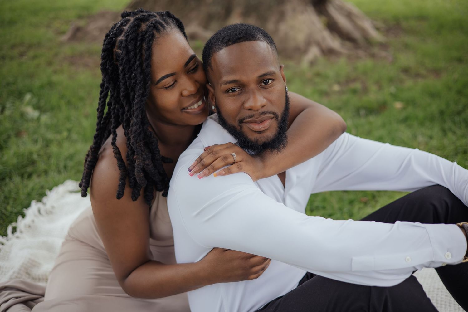 Black couple embracing on white blanket in park, woman hugging man from behind near tree trunk.