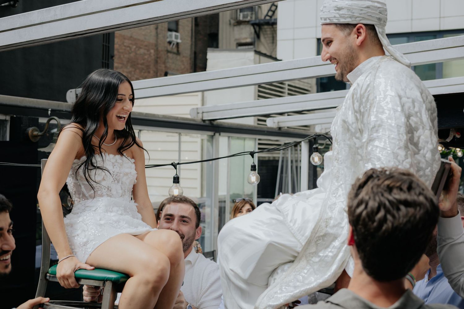 Bride and groom in traditional white attire are lifted on chairs during a joyful wedding celebration outdoors.
