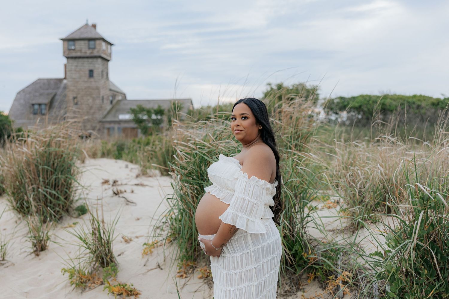 Pregnant woman in white off-shoulder gown poses on sandy beach dunes with historic stone building backdrop.