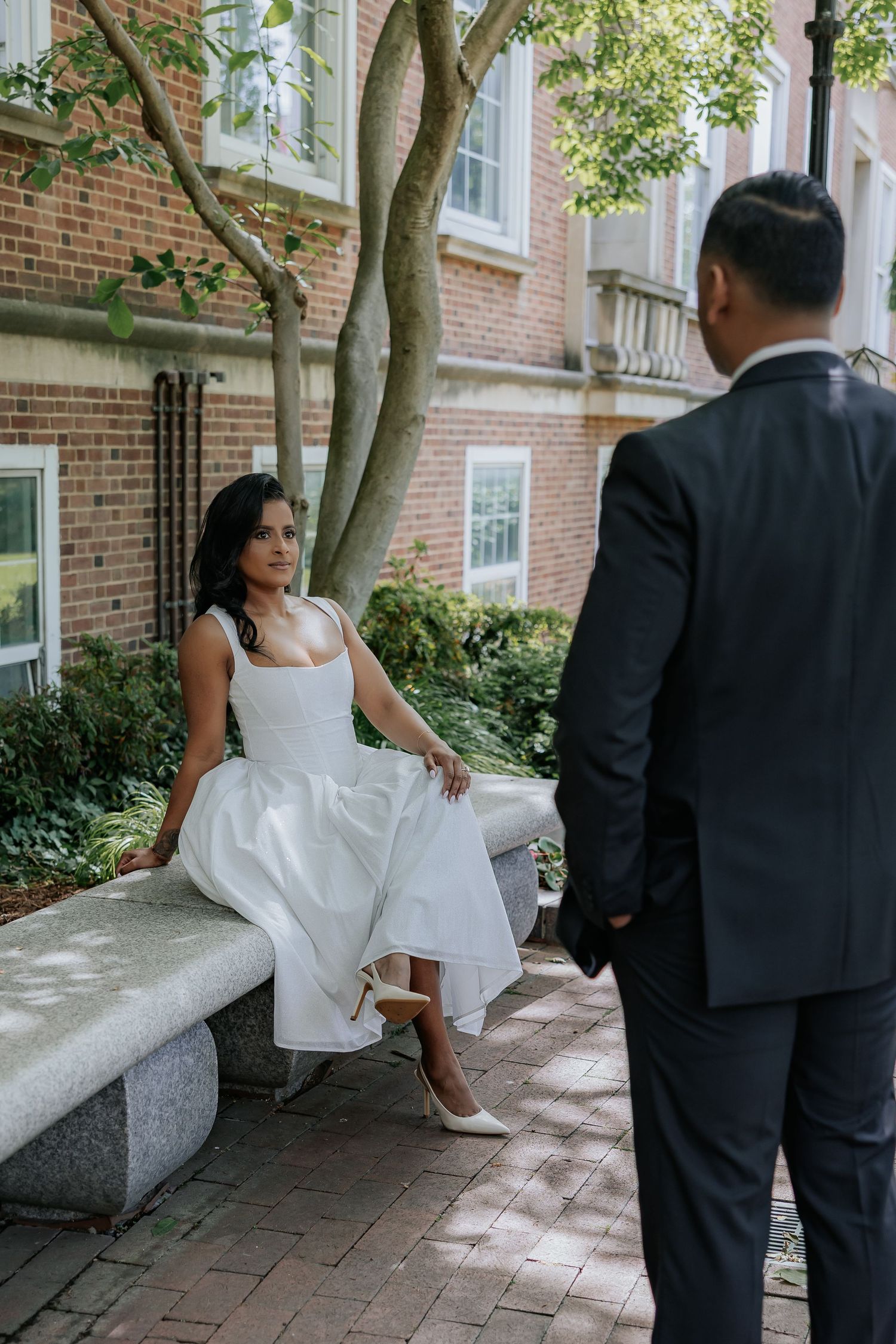 Elegant bride in tea-length white dress seated on stone bench as groom approaches in garden.