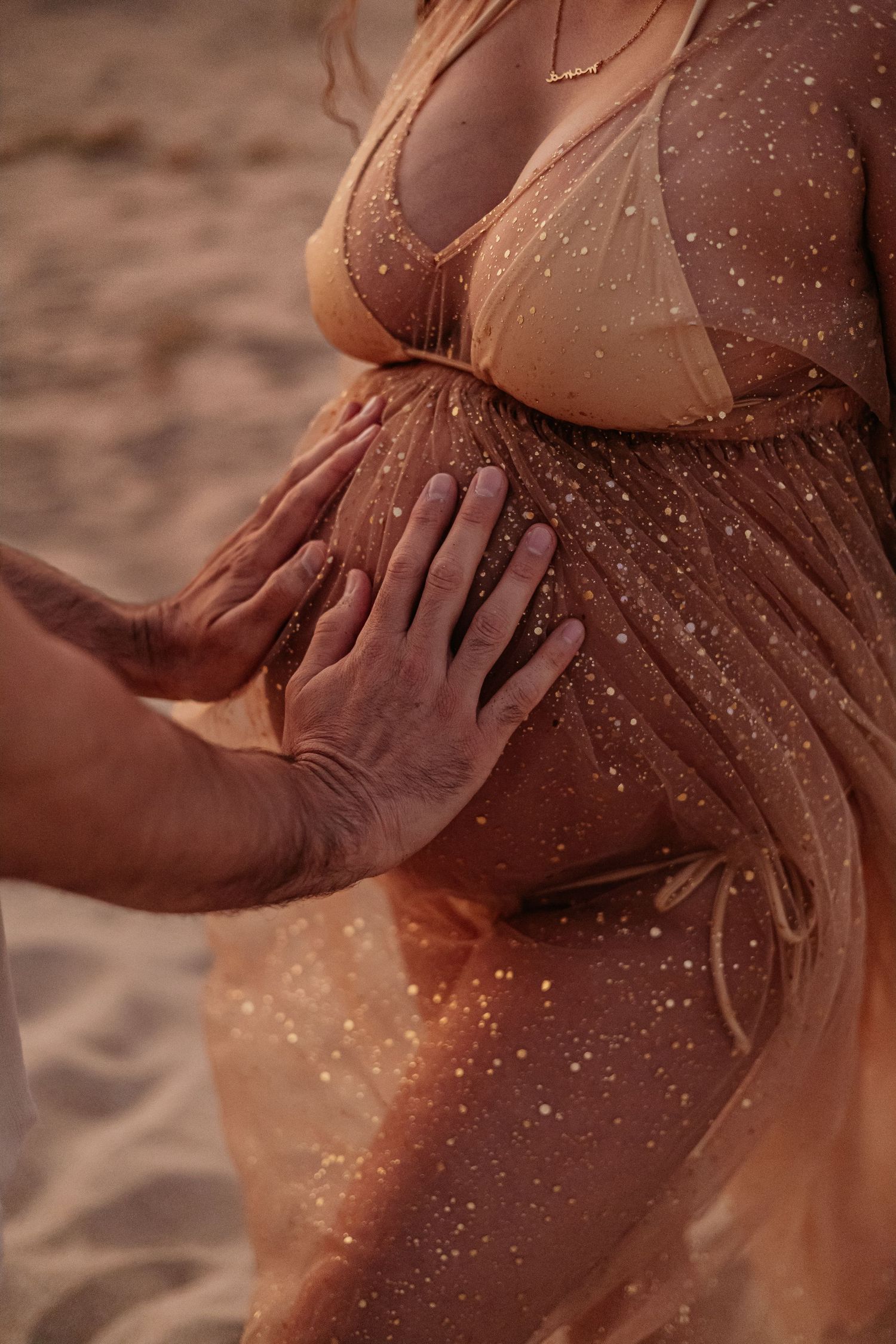 Close-up of hands cradling pregnant belly in sparkly gold tulle maternity beach photoshoot.