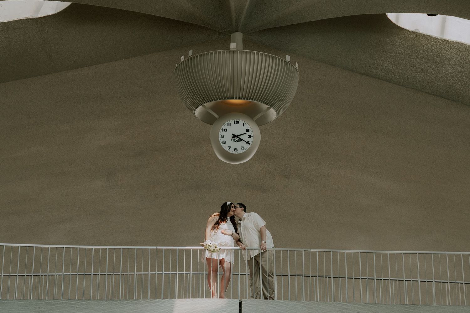 Couple shares romantic kiss on bridge under vintage clock at TWA Hotel terminal.