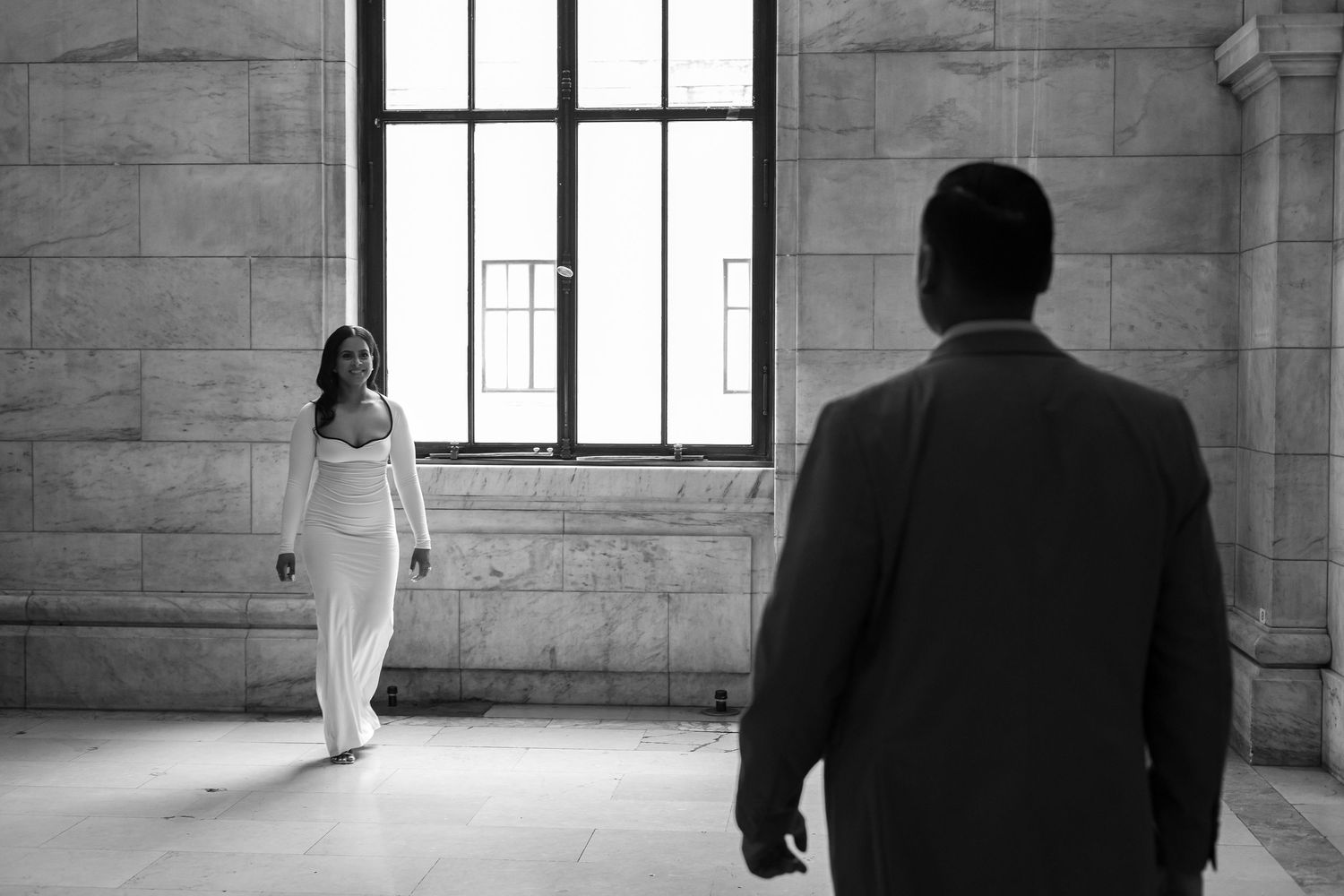 Black and white photo of a woman in white gown walking toward her partner.