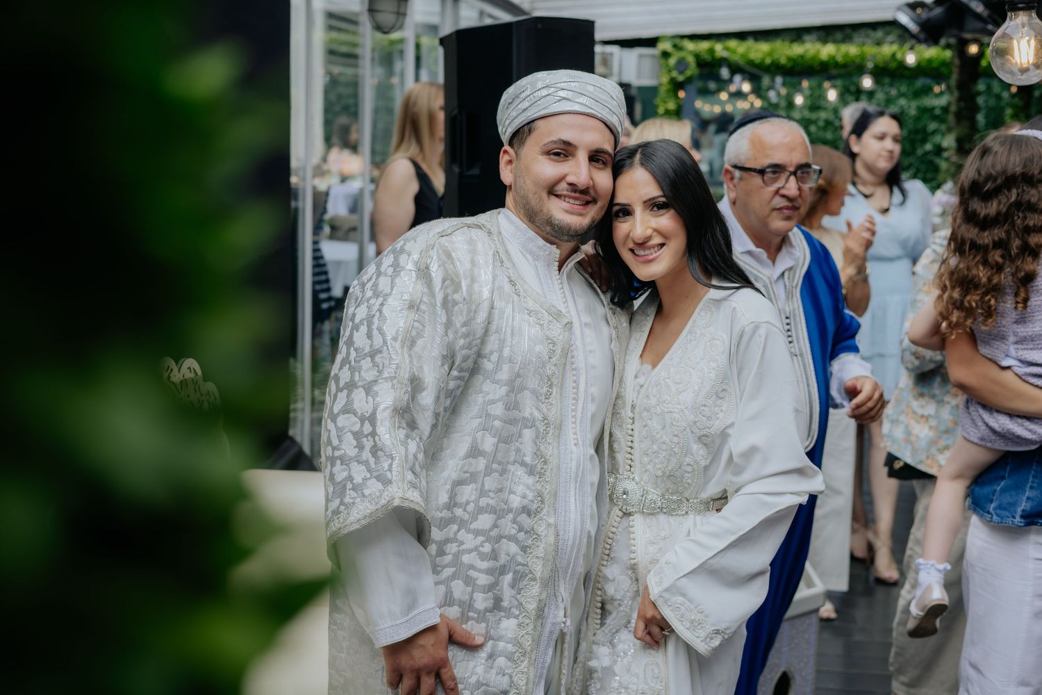 Couple in matching white traditional Moroccan wedding attire pose together smiling at outdoor celebration event.