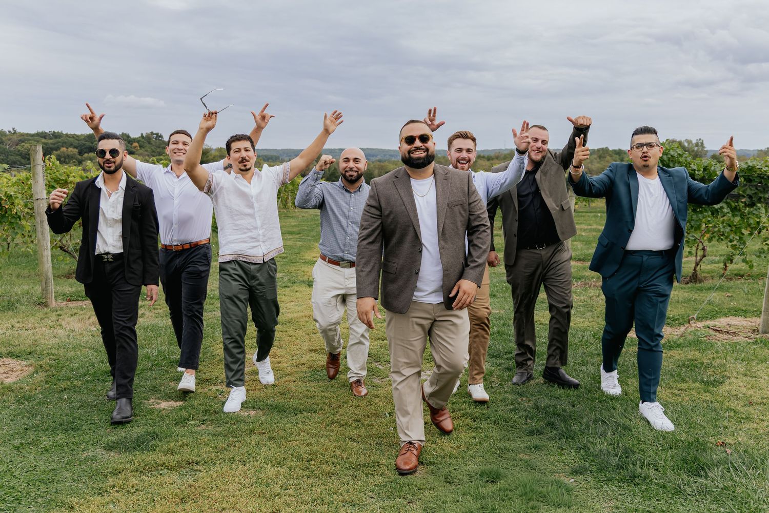 Seven men in semi-formal attire walking confidently and celebrating at an outdoor vineyard wedding.