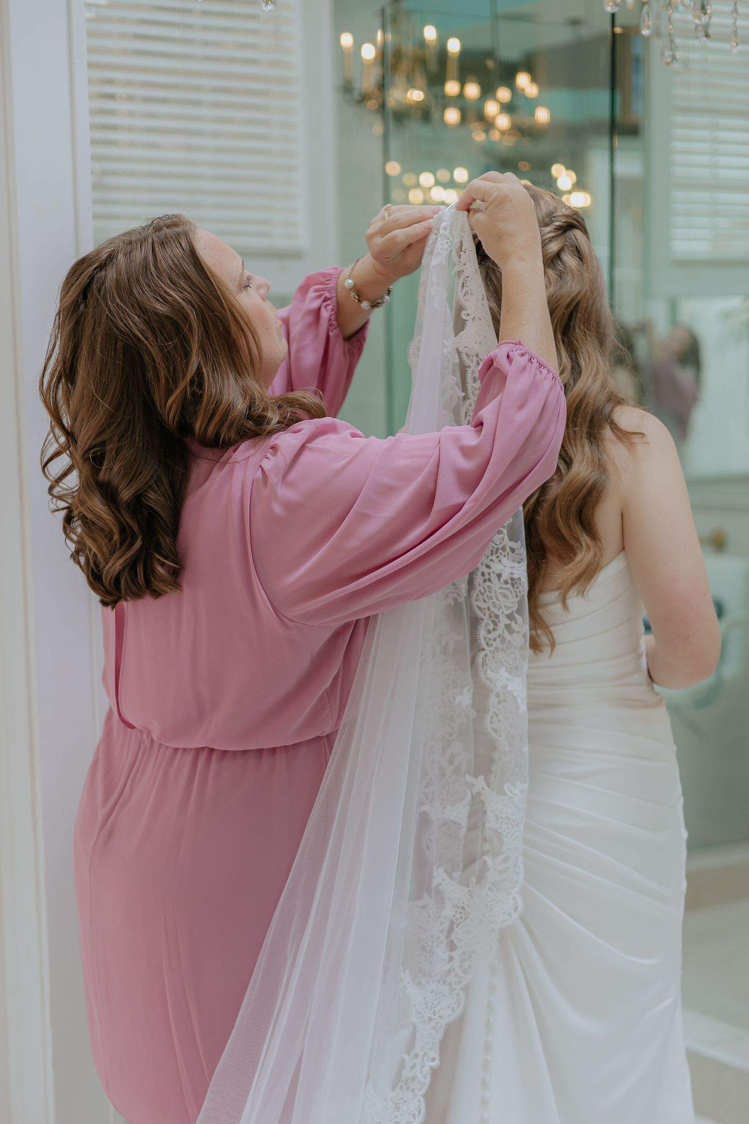 Woman in pink dress helping bride attach lace veil to her hair in bridal suite.