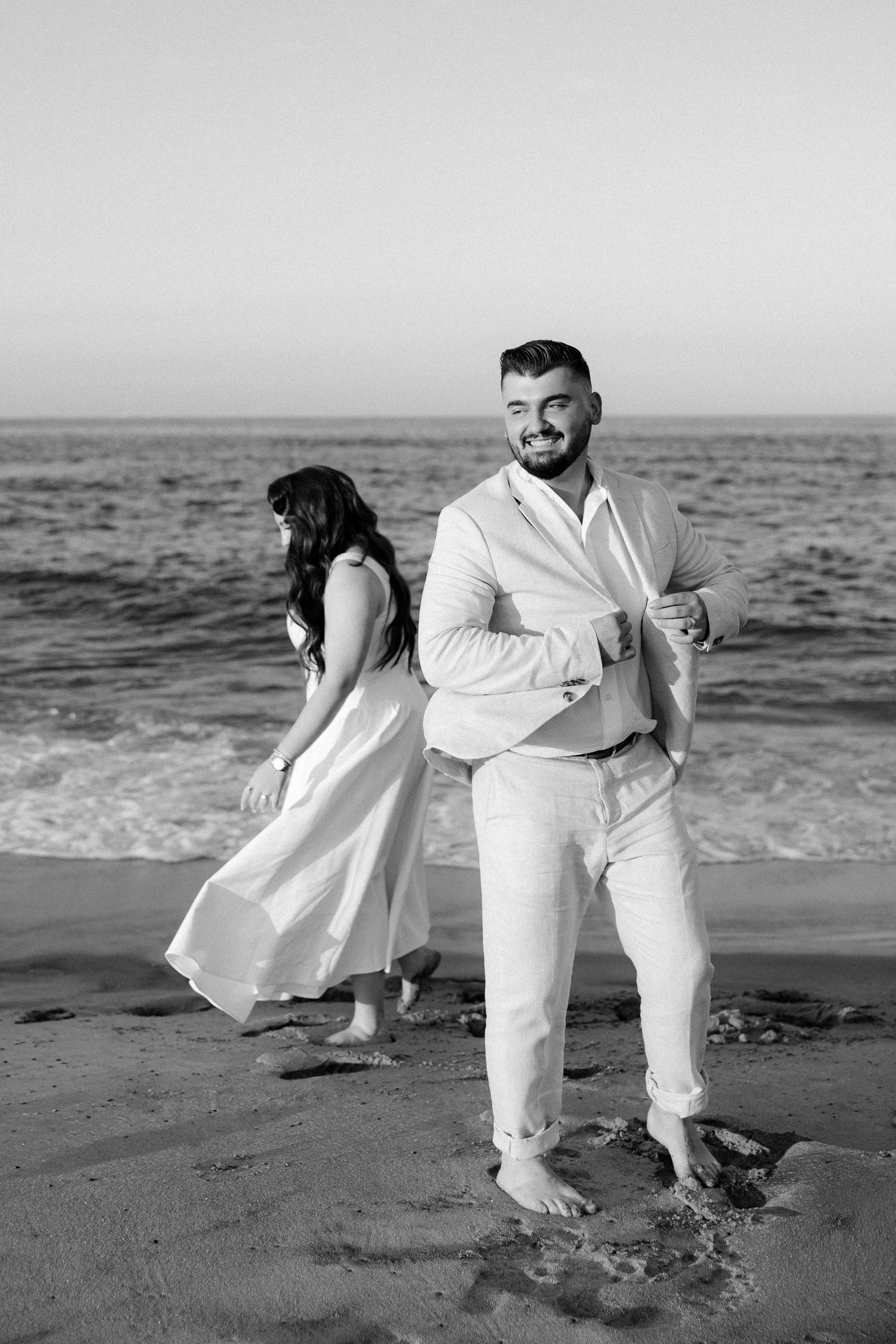 Black and white photo of a smiling groom in white suit on beach with bride in flowing dress by ocean waves.