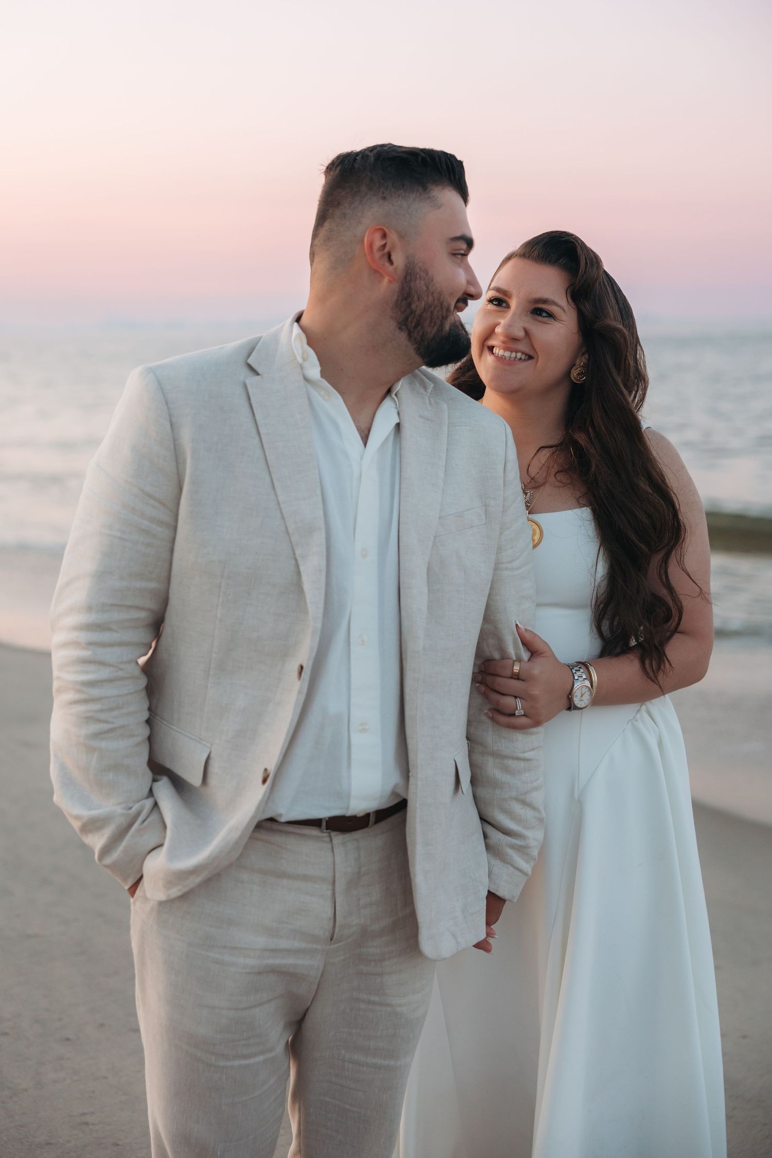 Couple smiling at each other on beach at pink sunset, man in linen suit, woman in white dress.