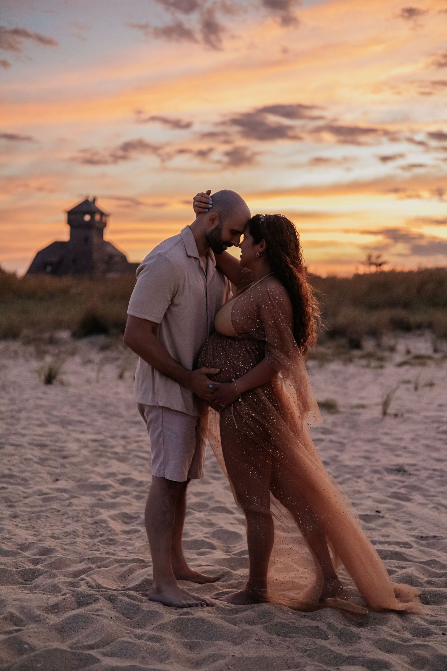 Couple sharing tender kiss while holding pregnant belly on beach during colorful sunset maternity shoot.