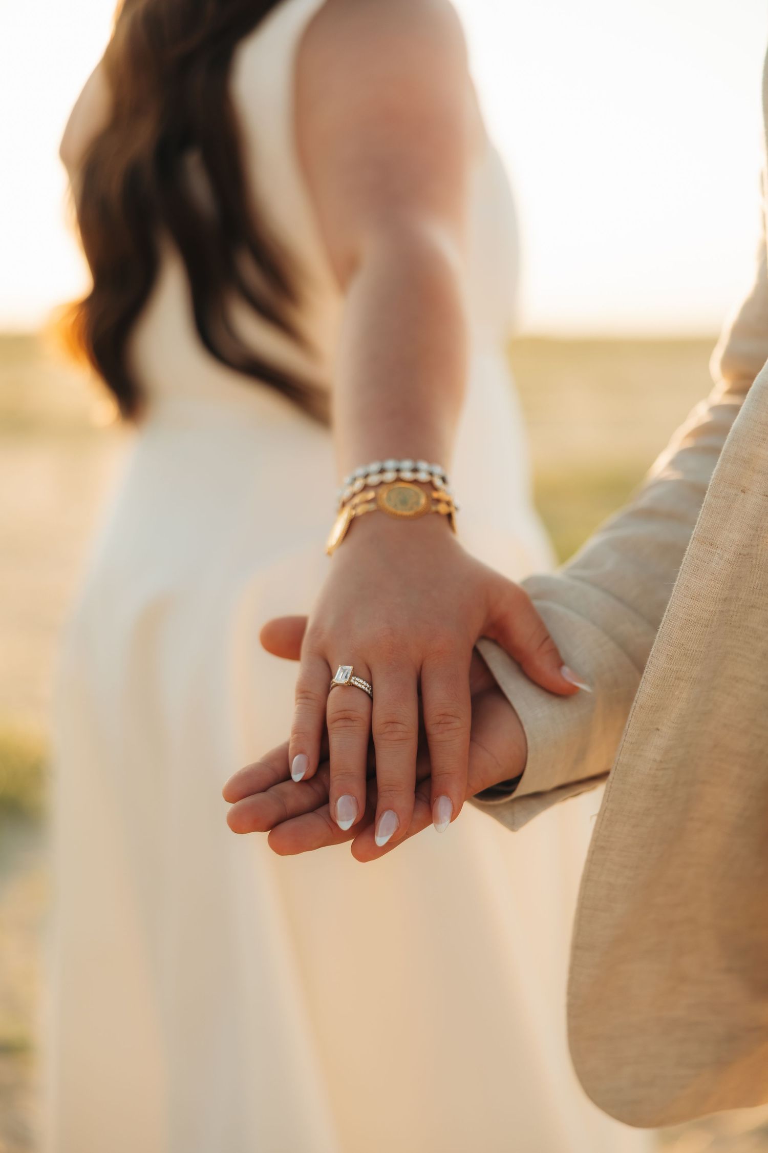 Close-up of couple holding hands at sunset, woman wearing diamond engagement ring and gold watch in white dress.