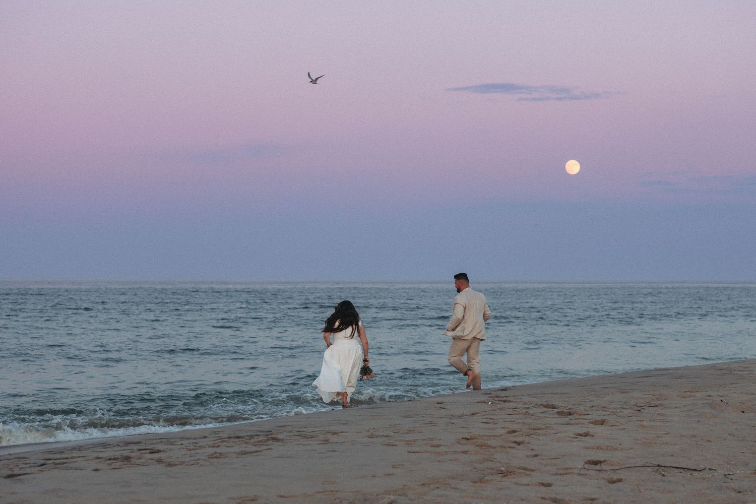Couple walking on beach at sunset with full moon rising over calm ocean waters in romantic twilight scene.