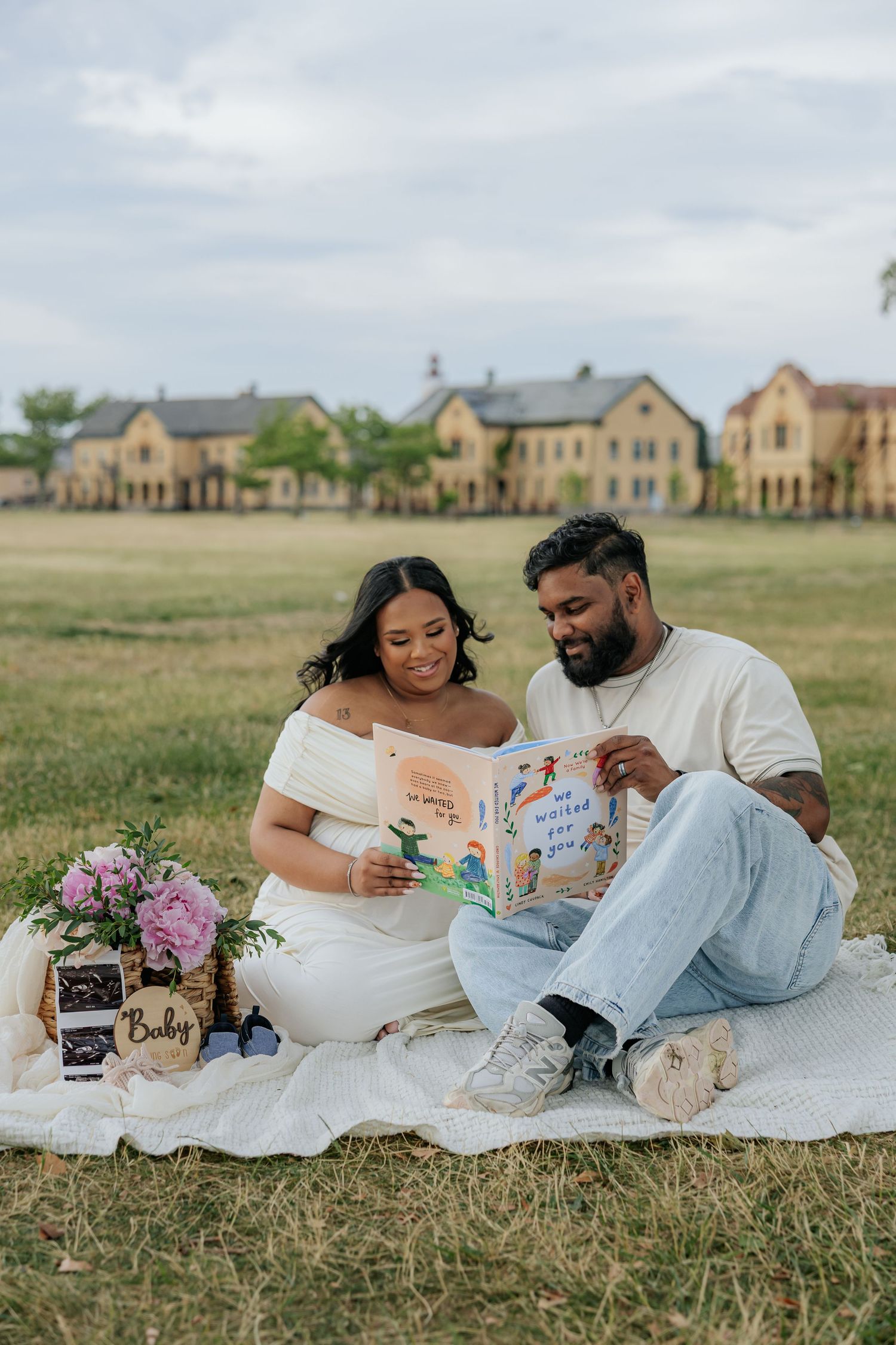 Couple sitting on blanket in field reading baby book with flowers and ultrasound photo during maternity photoshoot.