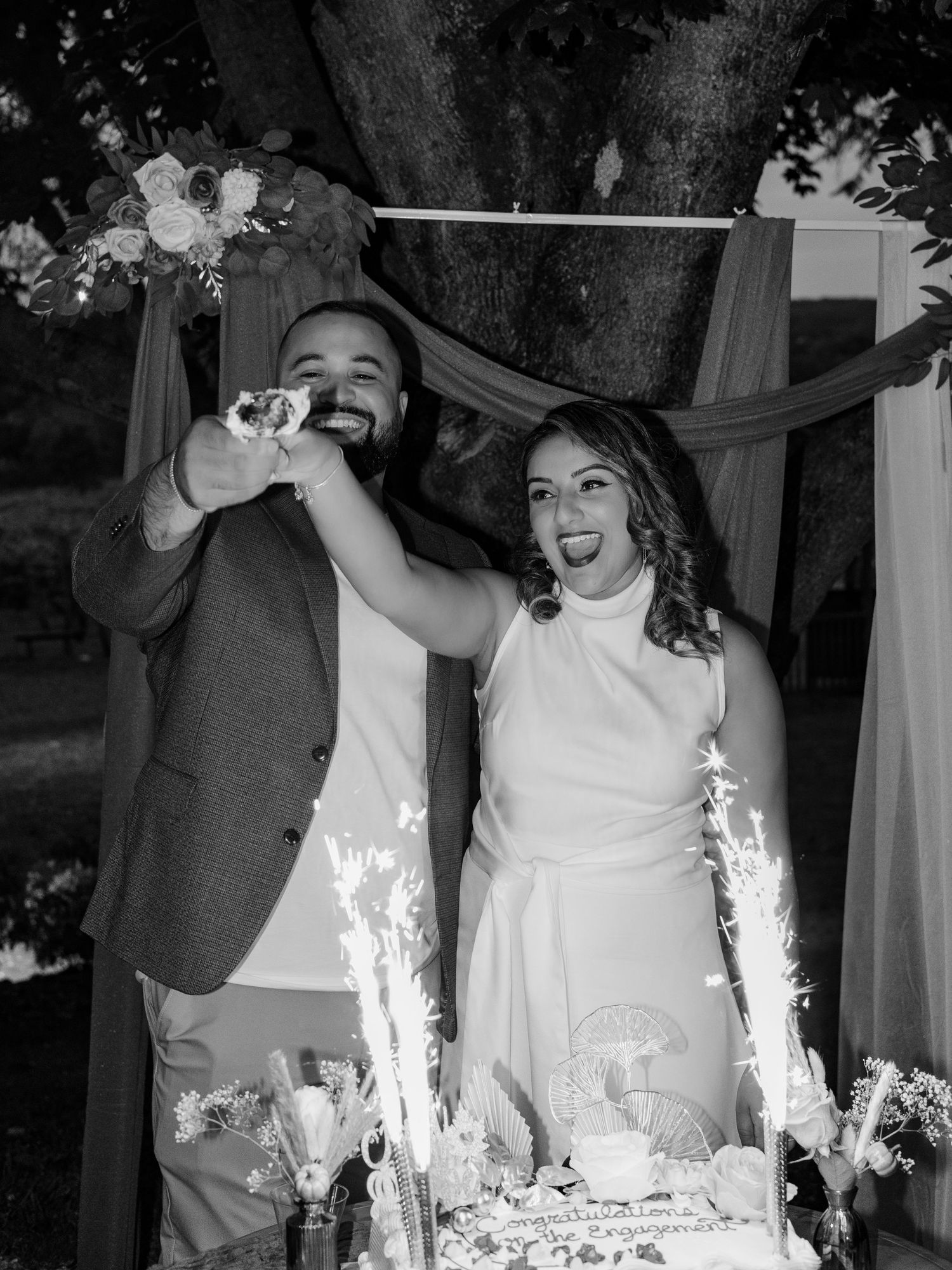 Black and white photo of couple laughing while feeding each other engagement cake.