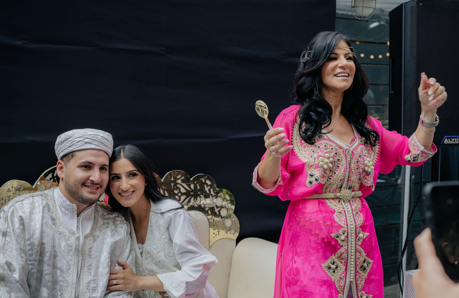 Woman in pink embroidered kaftan celebrates with spoon while couple in white traditional attire sit on wedding throne.