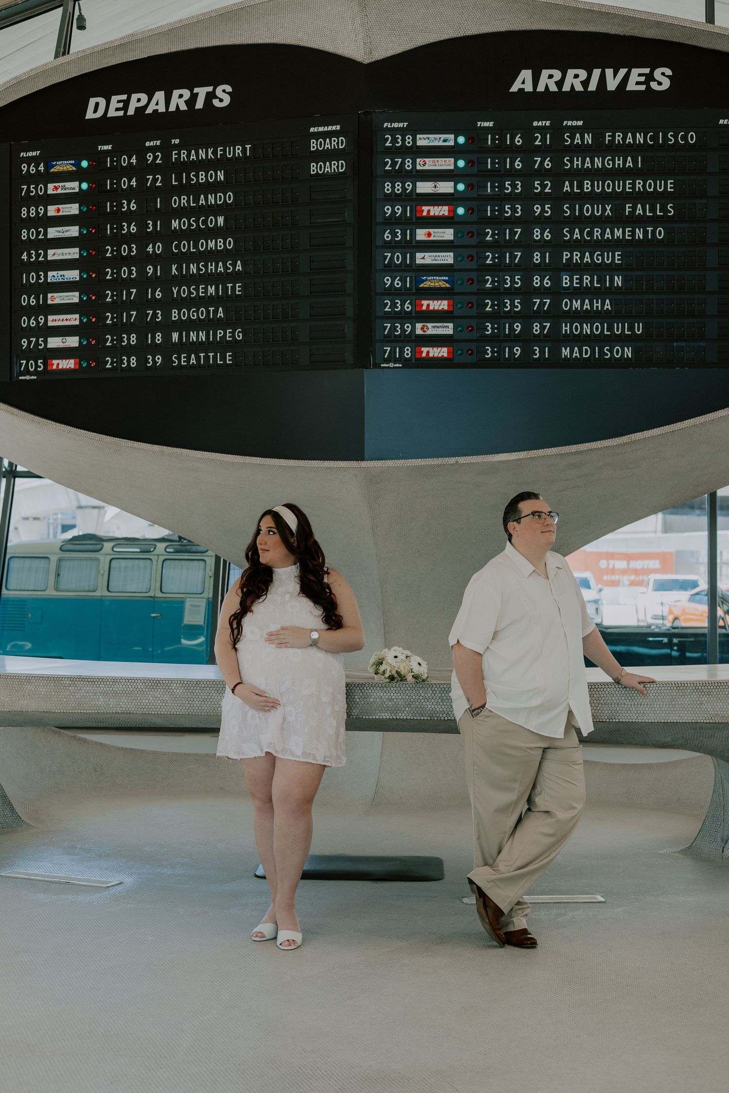 Expecting couple standing before a vintage airport departures and arrivals board at the TWA Hotel.