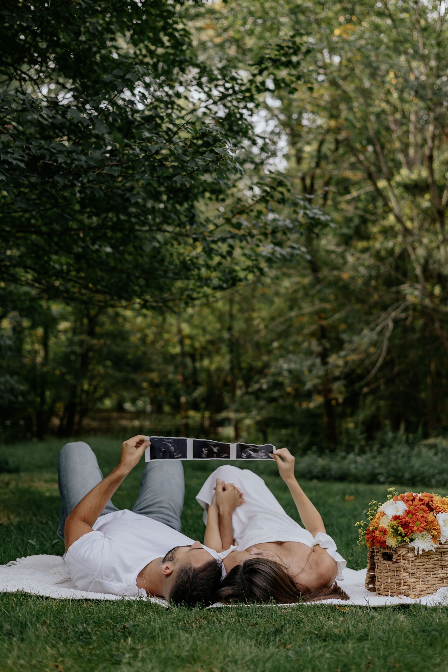 Couple lying on white blanket holding ultrasound photos above them with autumn floral arrangement nearby.