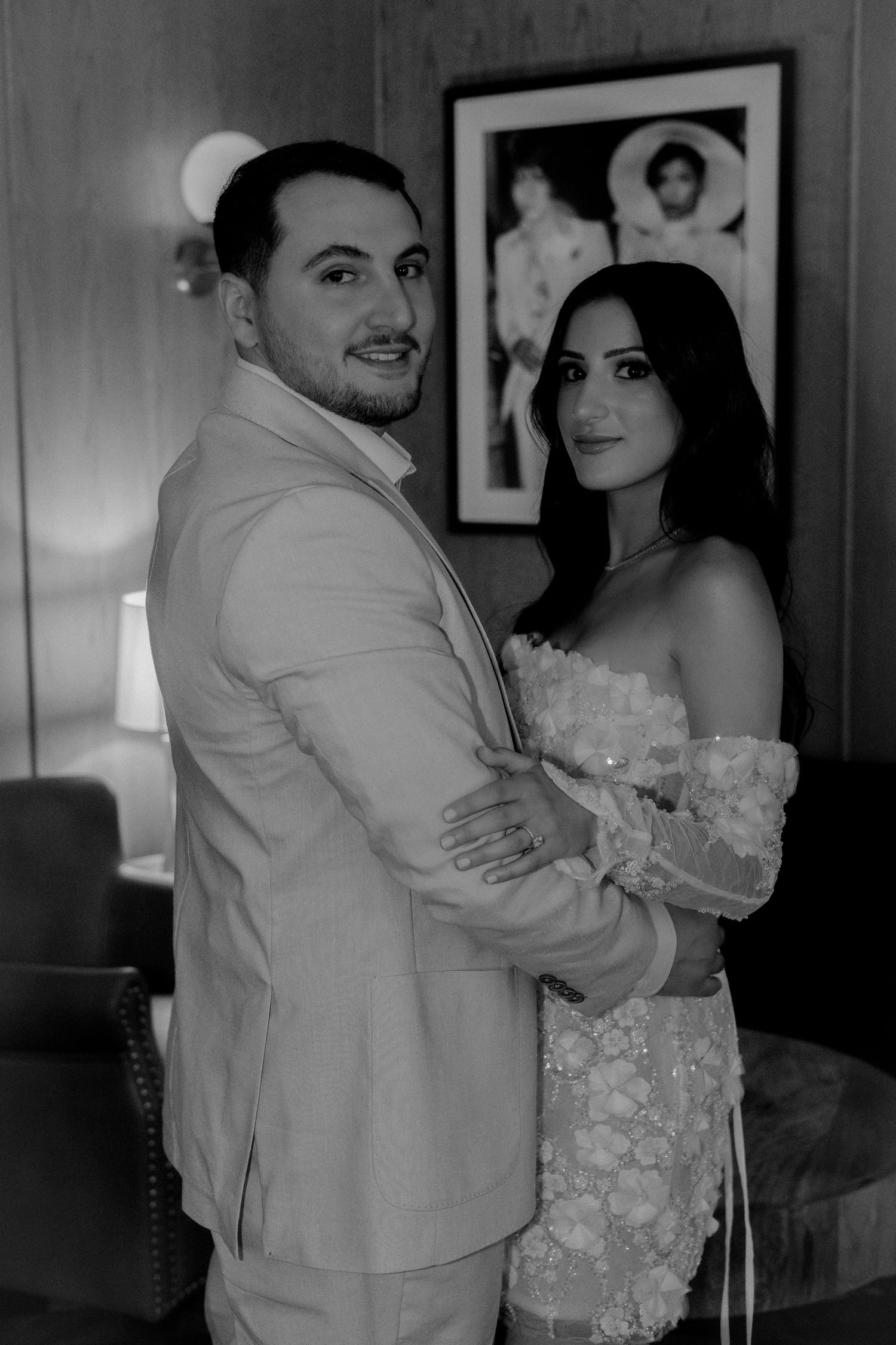 Black and white portrait of wedding couple embracing indoors with framed artwork behind them.