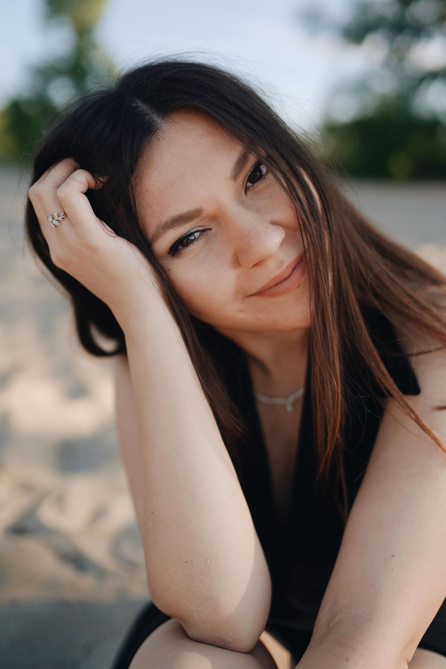 Close-up portrait of smiling brunette woman with blue eyes sitting on sandy beach at golden hour.