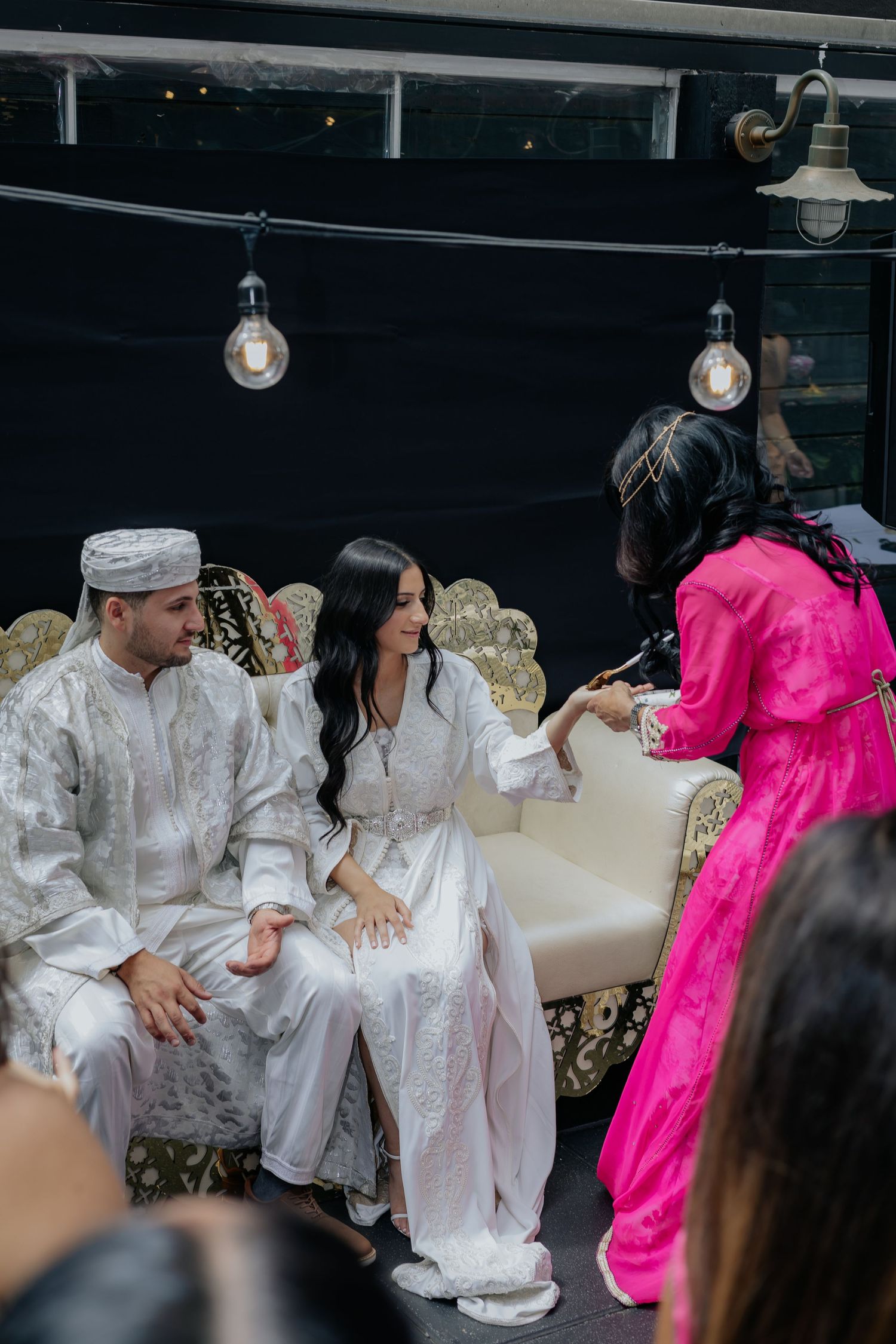 Woman in pink kaftan attends to bride and groom in white Moroccan attire seated on ornate gold wedding sofa.