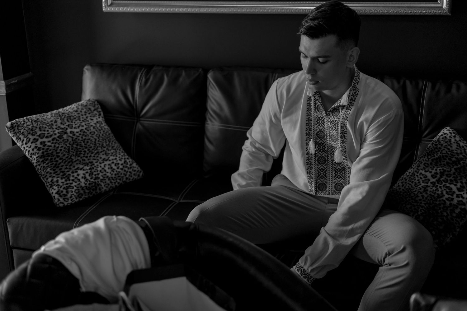Black and white photo of groom in embroidered shirt sitting on leather sofa preparing.