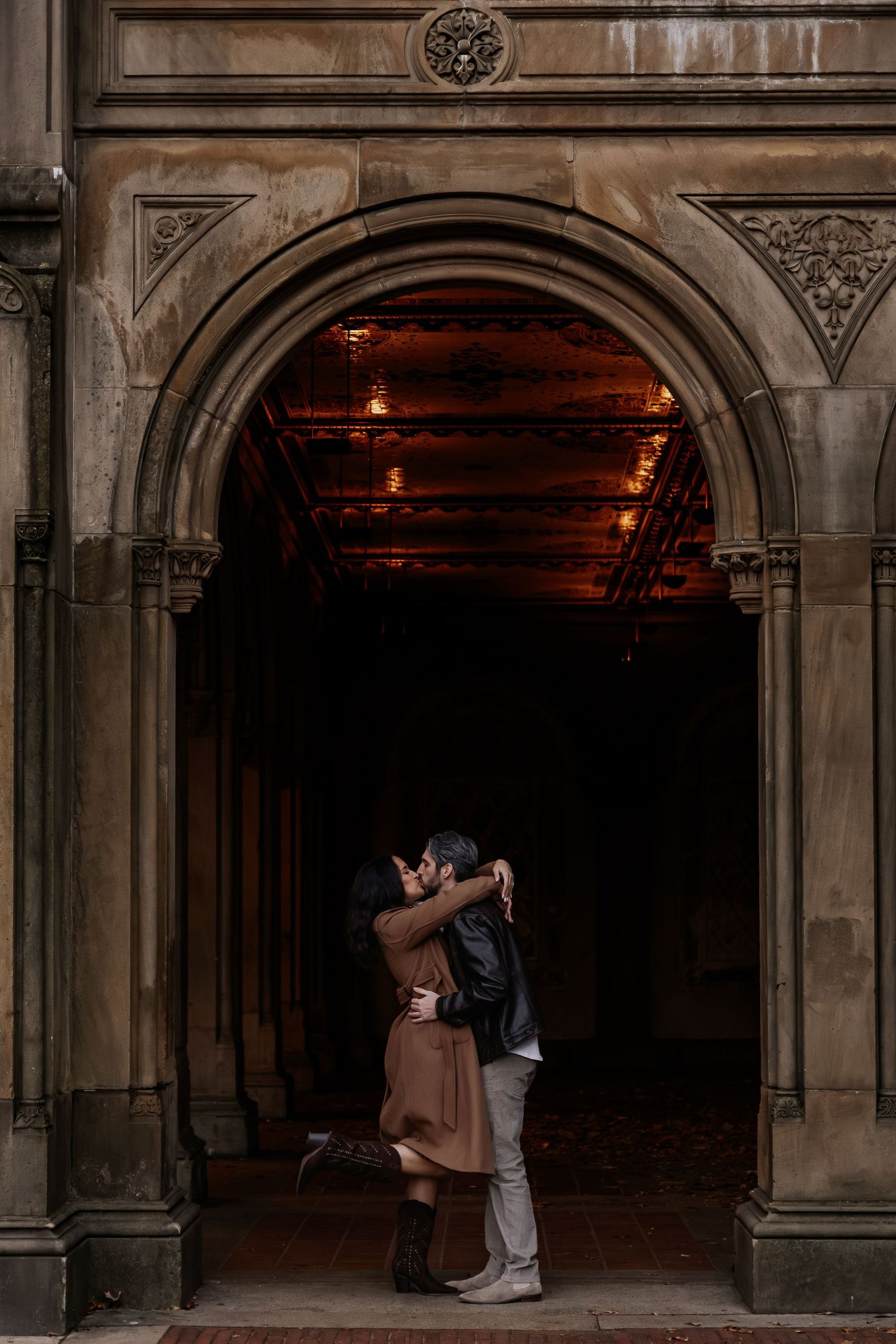 Couple kissing under a grand stone arch with warm glowing lights in the background.