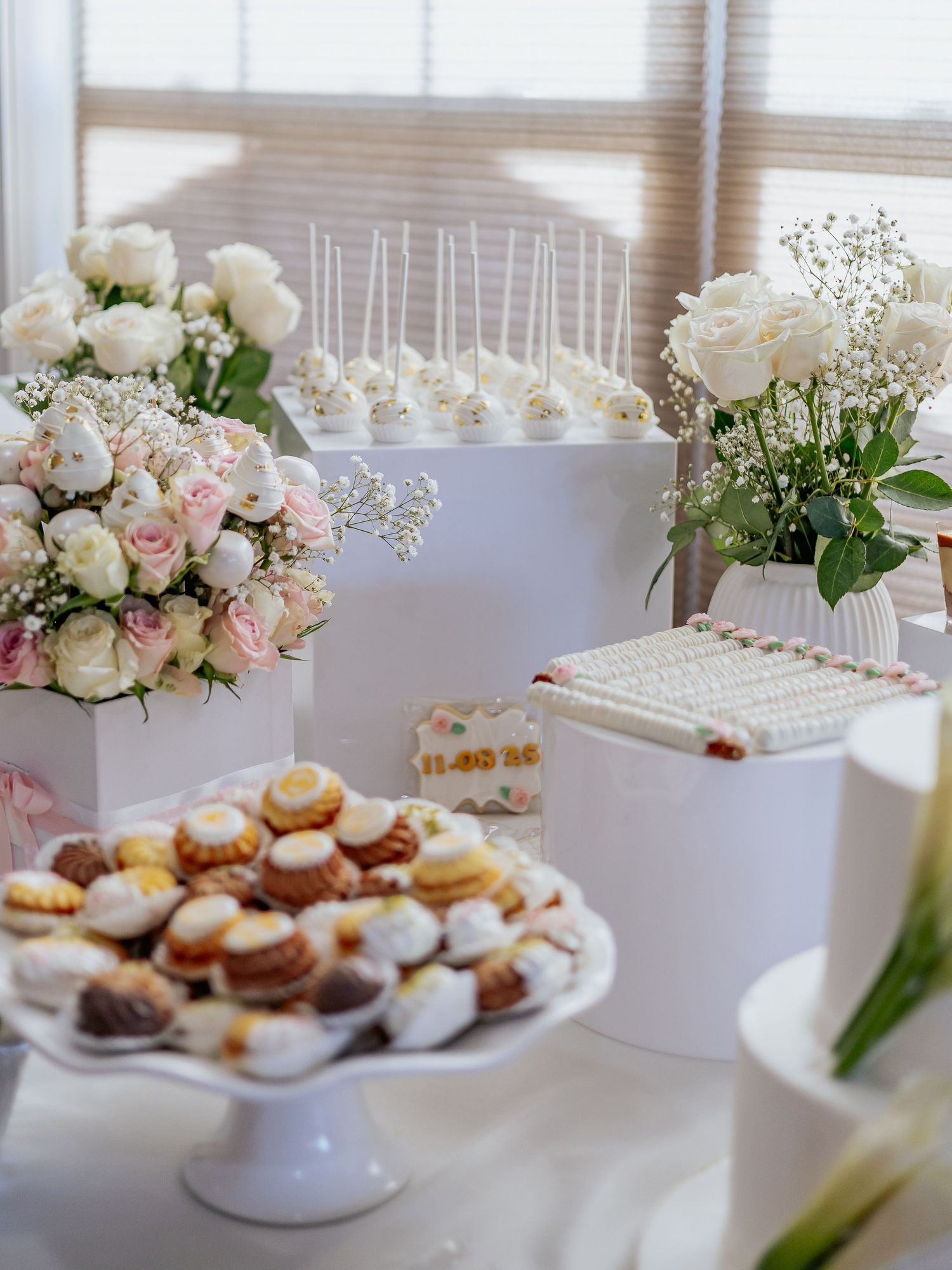 Elegant wedding dessert table with white cake pops, macarons, and floral arrangements of white and pink roses.