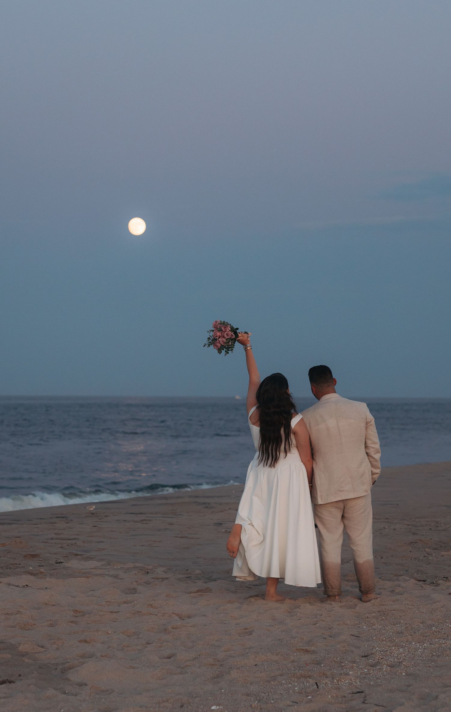 Couple in wedding attire standing on beach at dusk, bride raising bouquet toward full moon over ocean.