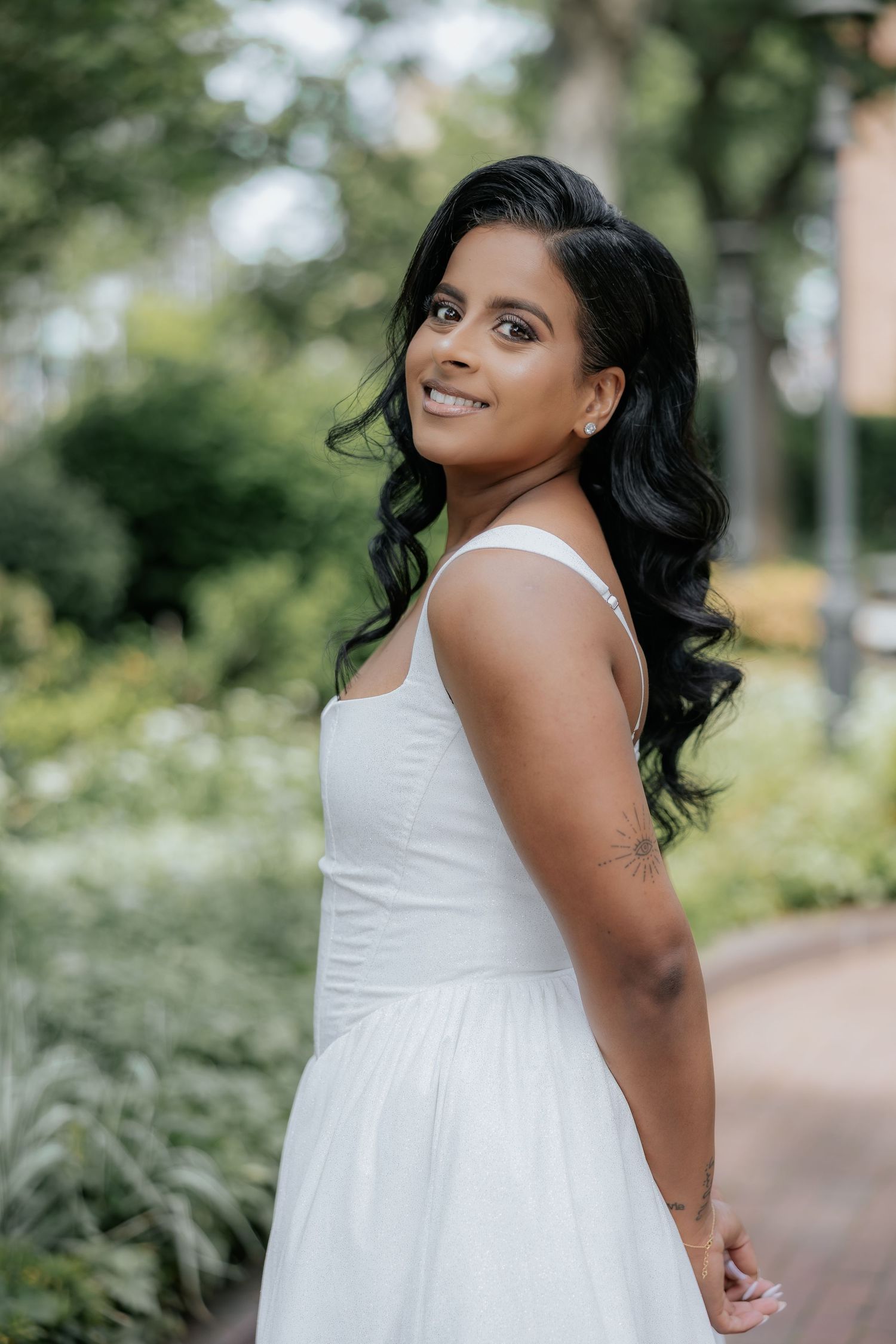 Beautiful South Asian bride in white dress smiling over shoulder in garden setting with lush greenery.
