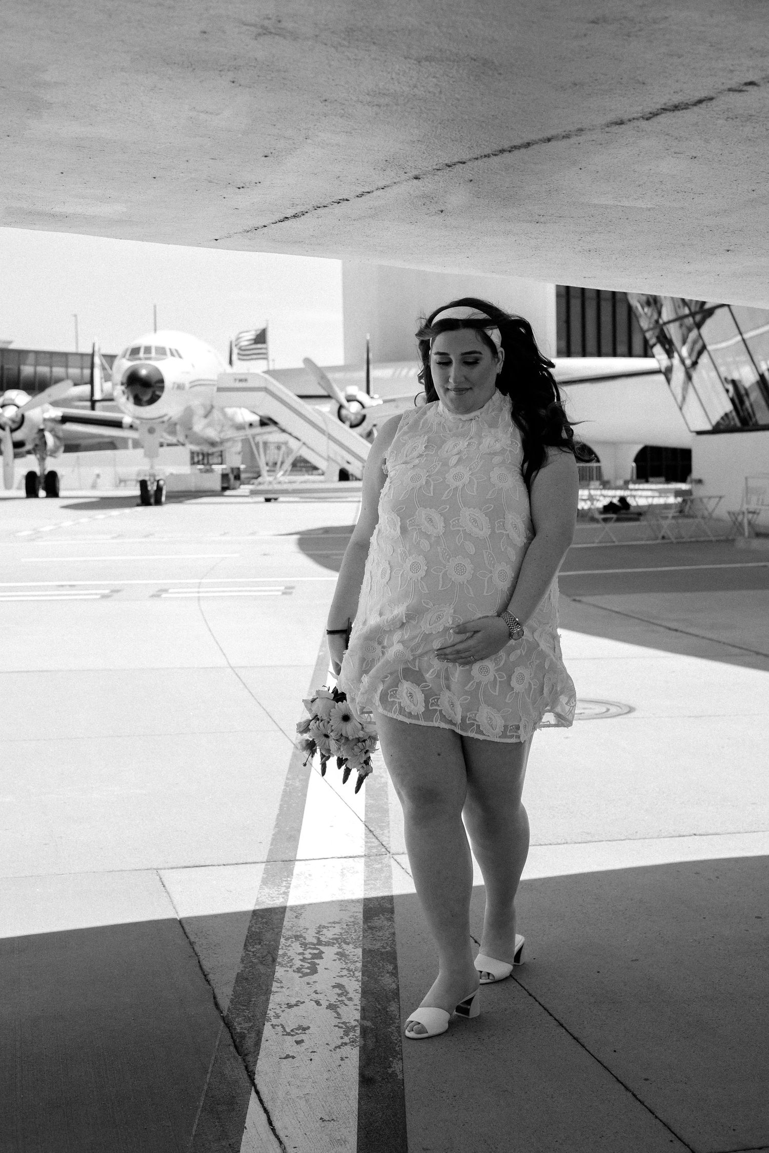 Pregnant bride in white dress holds bouquet on airport tarmac with vintage aircraft in background.