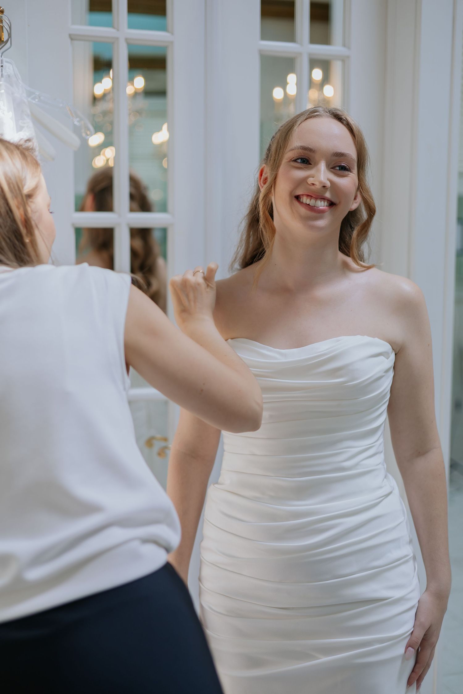 Smiling bride in strapless white wedding gown being assisted by a bridal consultant in a boutique.