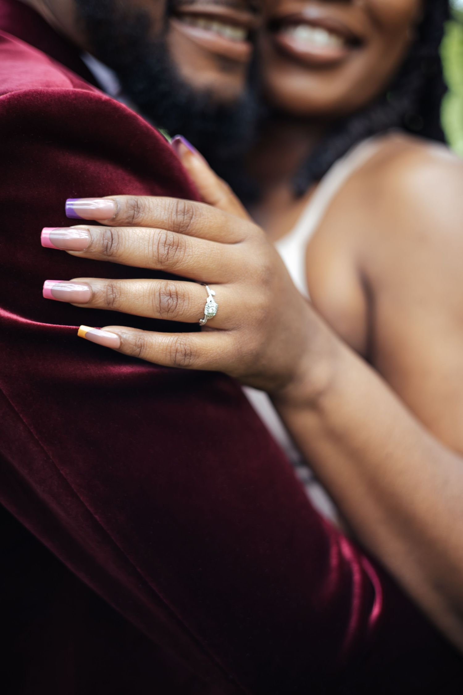 Close-up of bride showing diamond engagement ring with colorful ombre nails embracing groom in red velvet blazer.