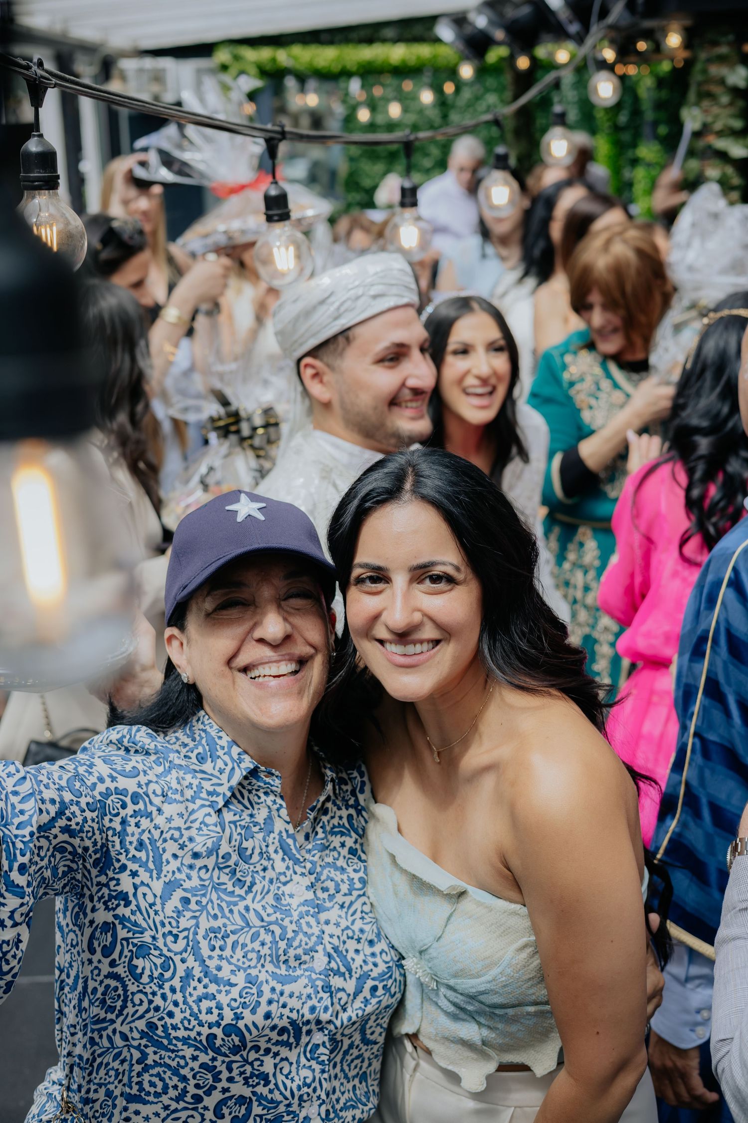 Two smiling women take a selfie at an outdoor wedding celebration with festive string lights and guests in background.