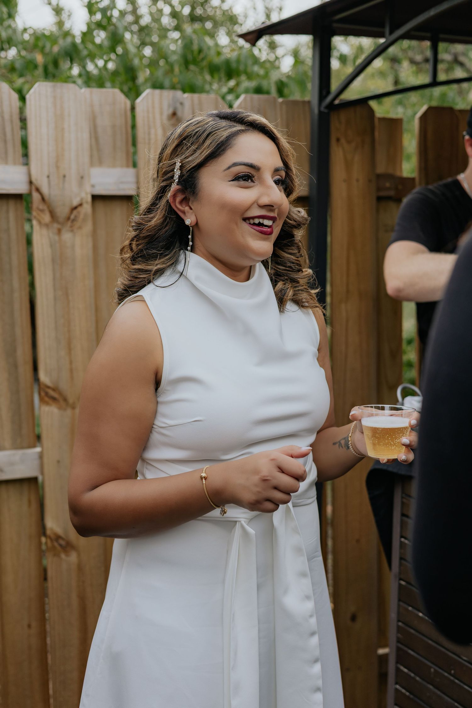 Bride in white sleeveless dress smiling while holding drink at outdoor engagement party.