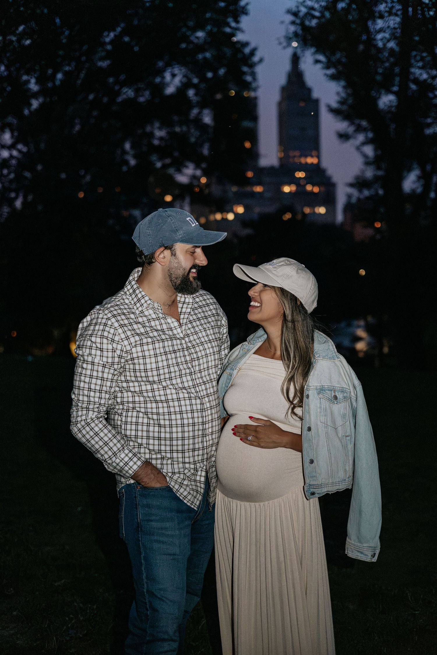 Smiling expecting couple in baseball caps with NYC city lights at dusk.