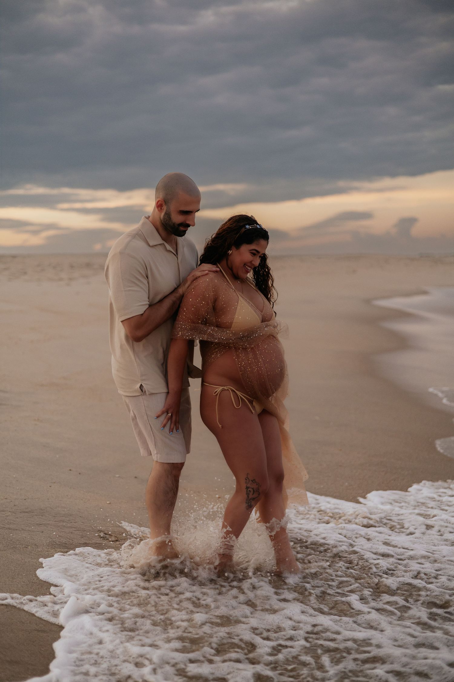 Expecting couple laughing in ocean waves at sunset, woman in gold sparkle bikini maternity shoot.